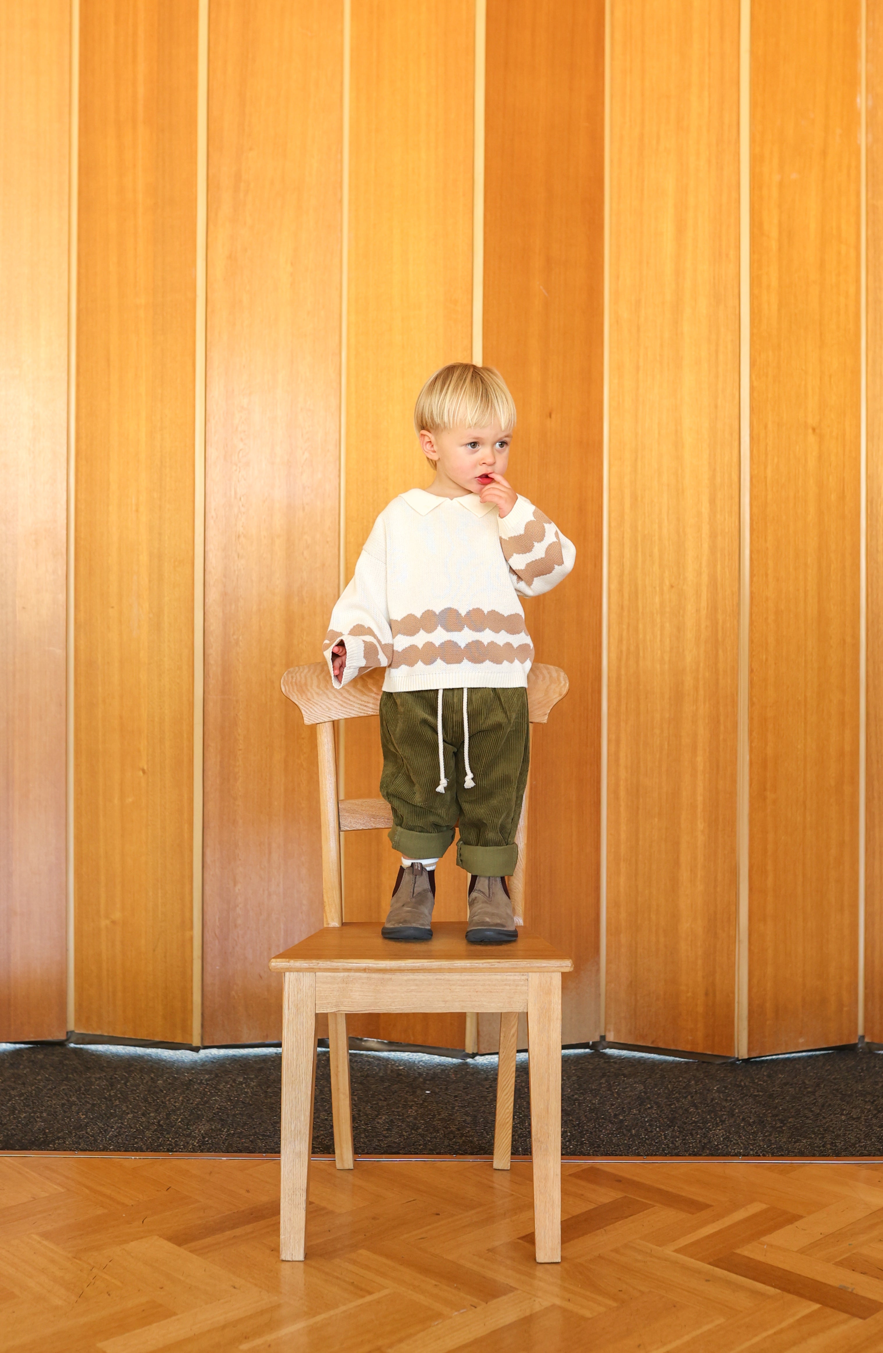 A blond child stands thoughtfully on a wooden chair, wearing a GROWN white sweater with a brown pattern and Grown ~ Organic Cord Pant Herb featuring an elastic waistband. The backdrop showcases wooden paneling, and the outfit is made from organic cotton with one hand touching their face.