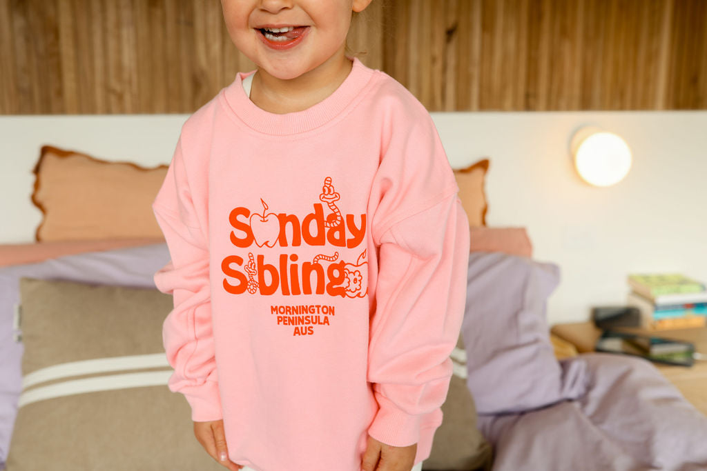 A young child wearing a high-quality Doc Sweater Pink by SUNDAY SIBLINGS, featuring "Sunday Sublime Mornington Peninsula AUS" printed in orange, smiles while standing on a bed. The background shows a wooden headboard, pillows, a light, and books stacked on a bedside table.