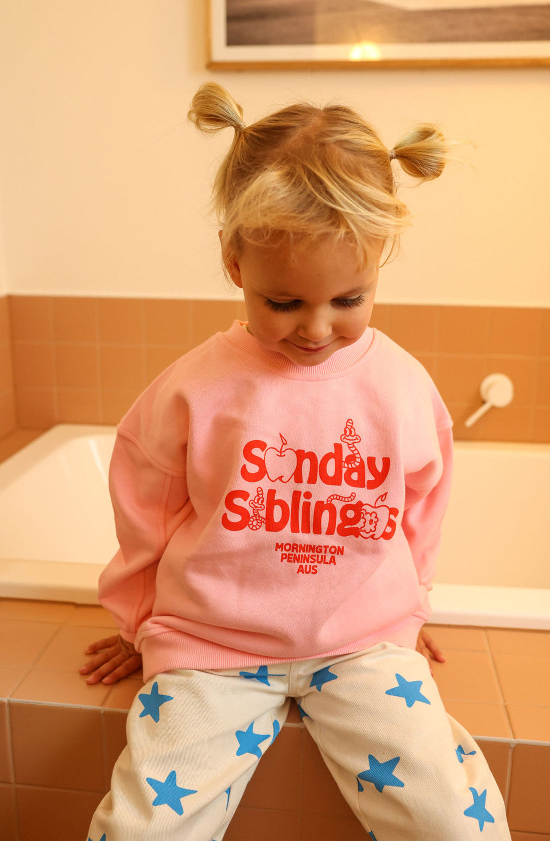 A child with blonde hair tied in two pigtails sits on the edge of a bathtub. The child is wearing a relaxed oversized fit Doc Sweater Pink from SUNDAY SIBLINGS, made from high-quality French terry cotton and emblazoned with red lettering that reads "Sunday Siblings" and "Mornington Peninsula AUS," paired with white pants featuring blue stars.