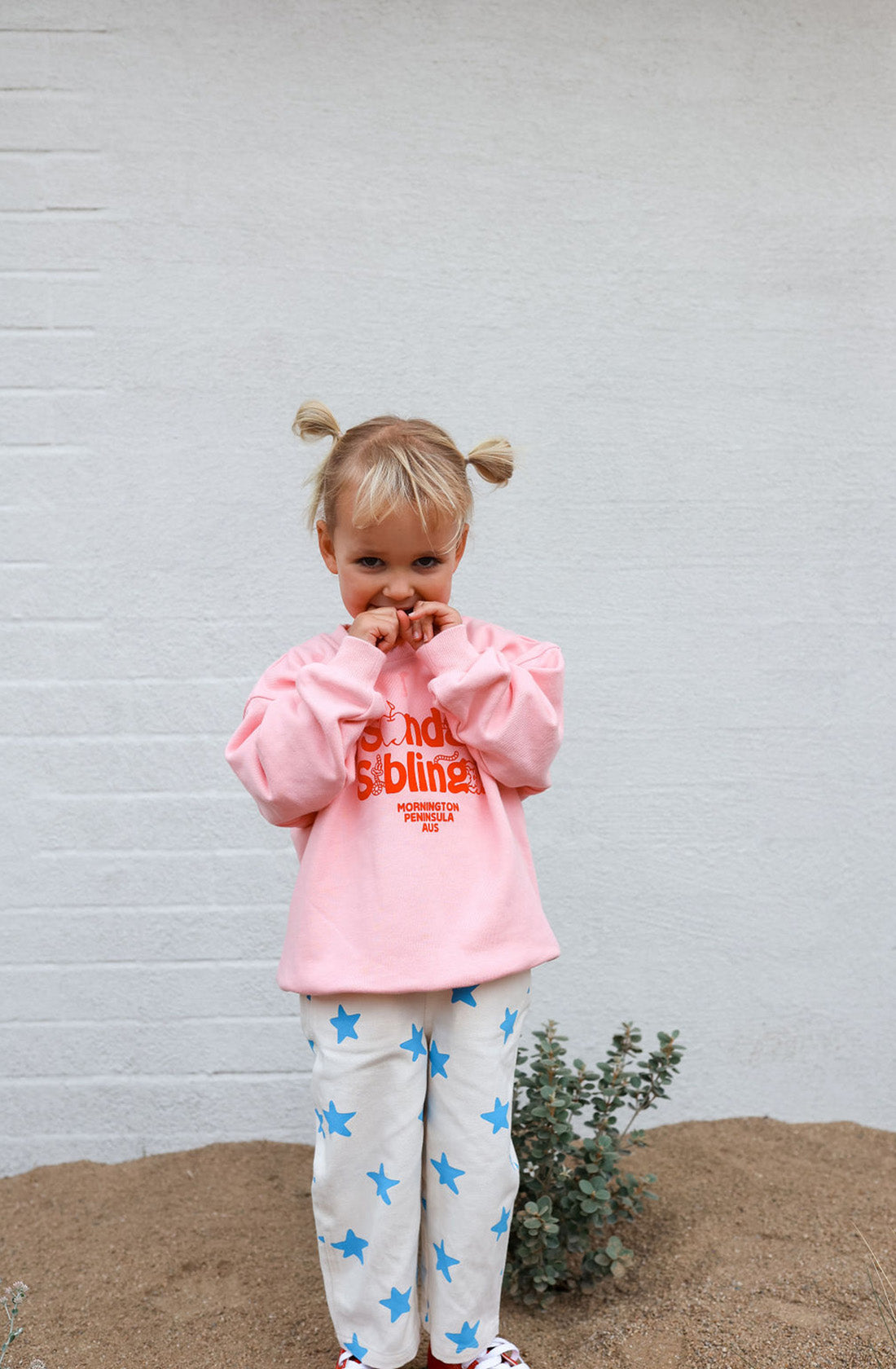 A young child with blonde hair styled in two buns stands against a white brick wall. They wear a pink Doc Sweater from SUNDAY SIBLINGS, crafted from high-quality French terry cotton, featuring red text and white pants adorned with blue stars. The child holds their hands near their face and appears to be smiling shyly.