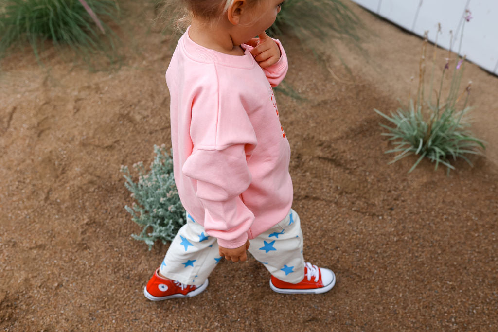 A small child wearing the Doc Sweater Pink by SUNDAY SIBLINGS in a relaxed oversized fit, white pants with blue stars, and red sneakers with cartoonish eyes on the toes walks outdoors on a sandy surface, with green plants and dry grass scattered around.