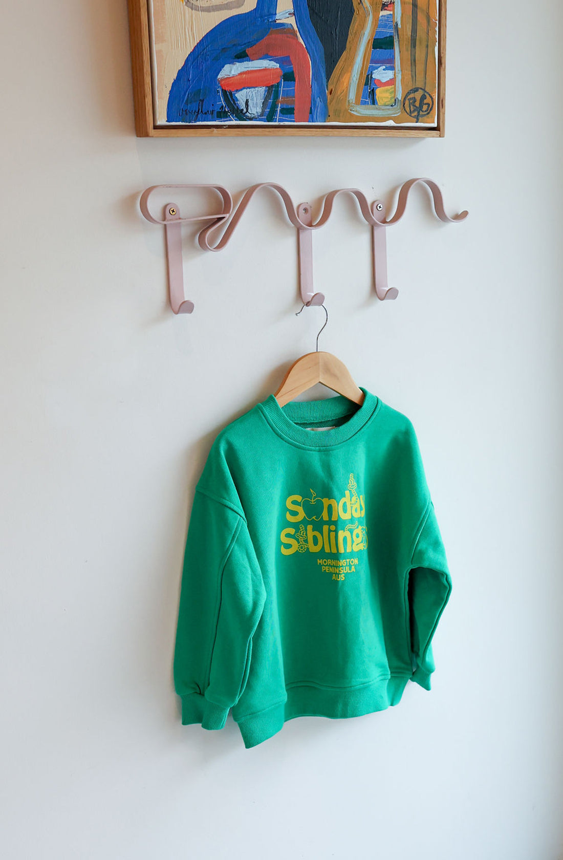 A Doc Sweater Green by SUNDAY SIBLINGS, crafted from soft French terry cotton and adorned with yellow text, hangs on a wooden hanger. The hanger is placed on a pink wall-mounted hook rack with five hooks. Above the sweater is a framed abstract painting by Doc Wilson Illustrations, featuring vibrant colors.