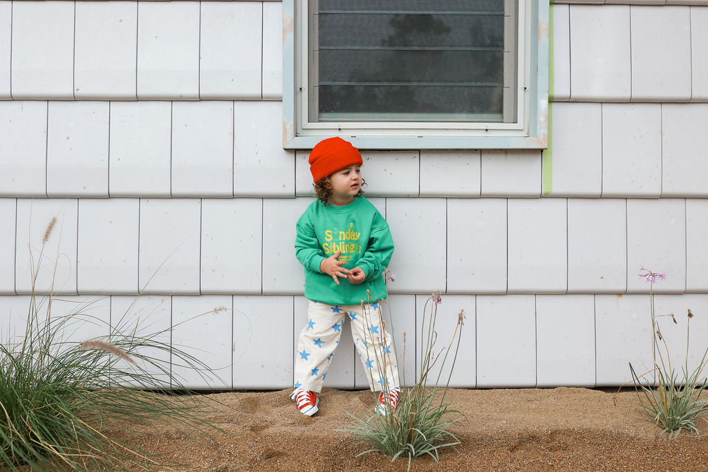 A small child in an oversized fit—a bright orange beanie, the "Doc Sweater Green" by SUNDAY SIBLINGS, and star-printed pajama pants made from cozy French terry cotton—stands outside in front of a light grey shingled wall with a window. Tall grasses grow near the child, who looks off to the side with hands clasped.