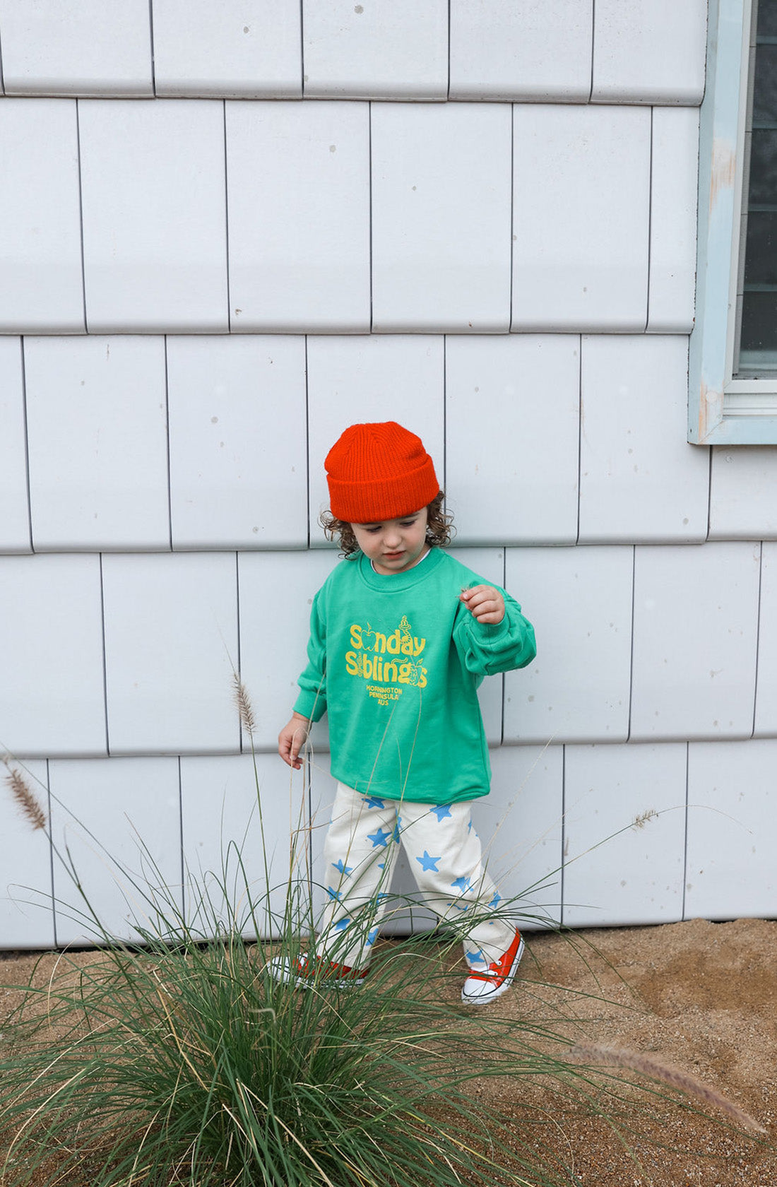 A young child stands against a white tiled wall, wearing the Doc Sweater in green made of French terry cotton by SUNDAY SIBLINGS, white pants with blue stars, red and white shoes, and an orange beanie. The child is reaching towards tall grass in front of them.