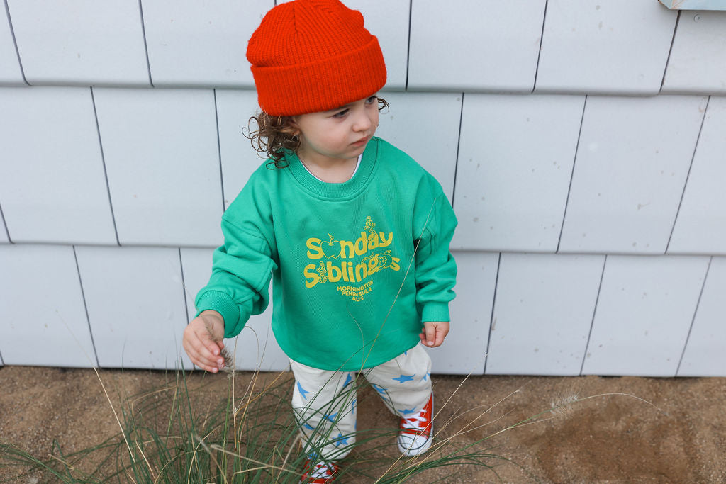 A young child stands outdoors wearing an oversized fit Doc Sweater Green by SUNDAY SIBLINGS, light-colored pants, and a vibrant orange beanie. Made of French terry cotton, their outfit is both stylish and comfortable. They have curly hair and are looking off to the side while touching a plant. The background consists of white shiplap siding.