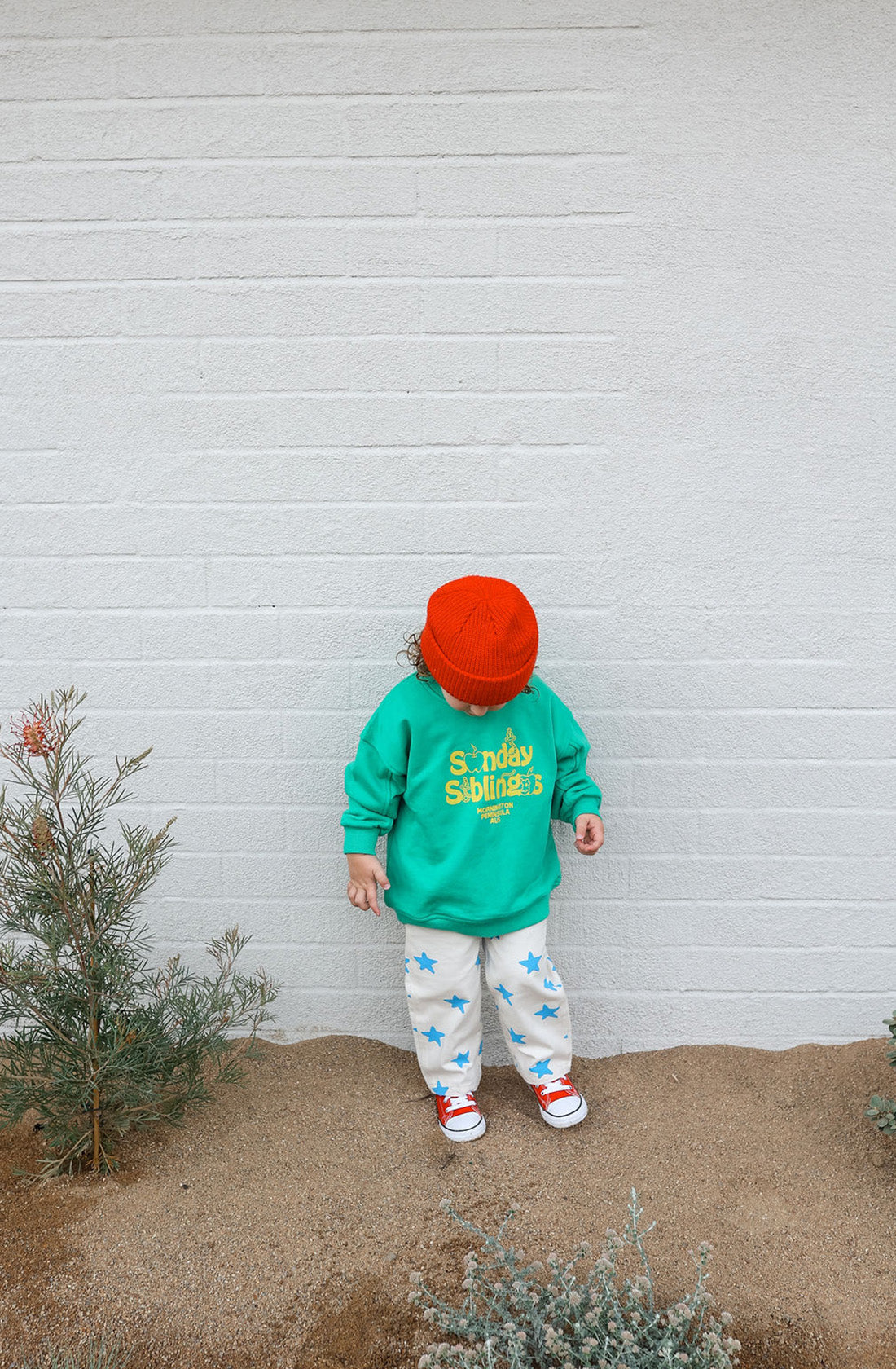 A child stands against a white brick wall, dressed in the SUNDAY SIBLINGS Doc Sweater Green, an oversized fit made of French terry cotton with text. The outfit is completed with white pants adorned with blue stars, red sneakers, and an orange beanie. The child looks down while two small shrubs are seen in front of the wall.