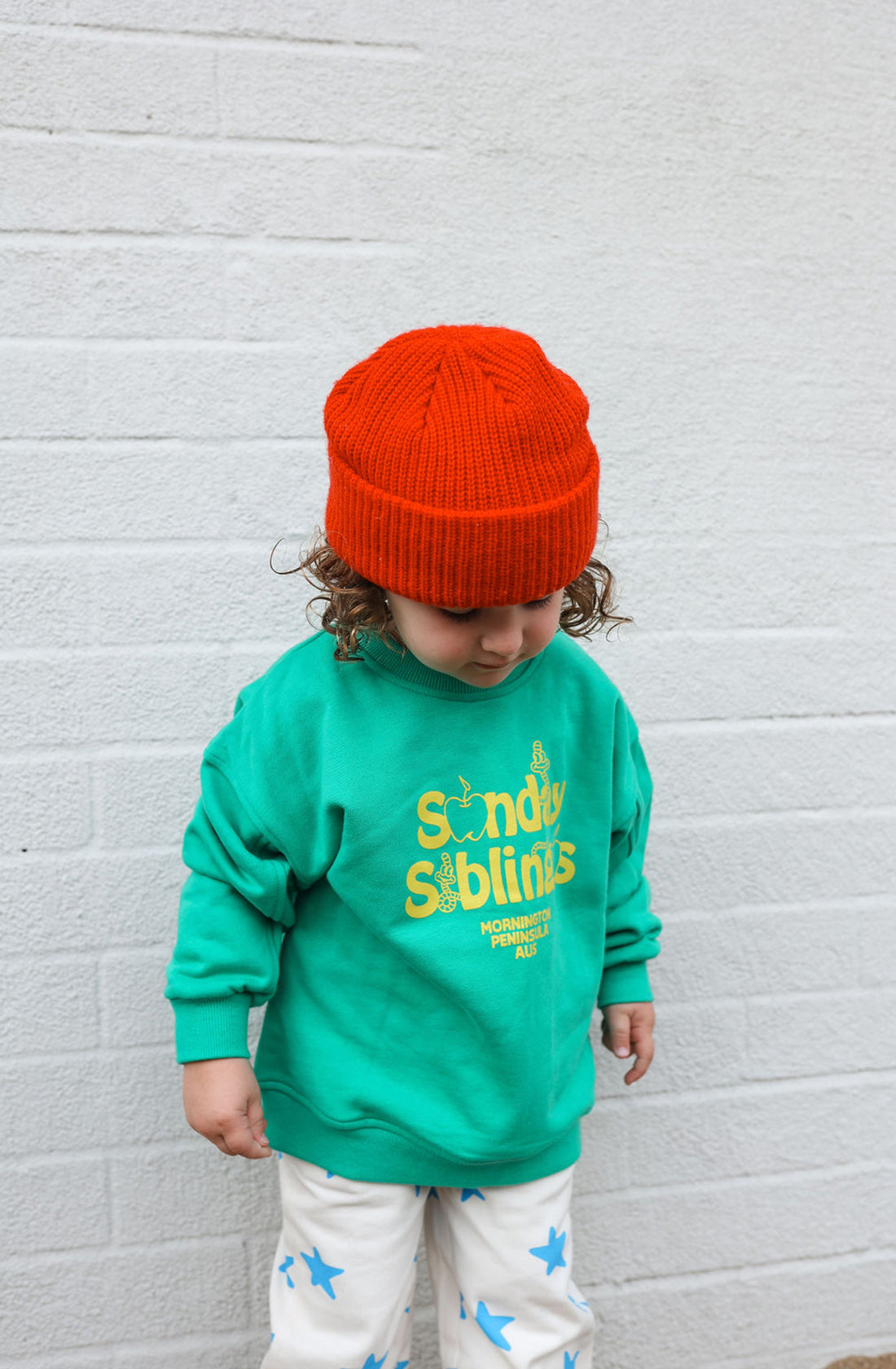 A toddler with curly hair, wearing a bright orange beanie, an oversized Doc Sweater Green by SUNDAY SIBLINGS made of soft french terry cotton featuring a colorful graphic design by Doc Wilson Illustrations, and white pants with blue stars, stands against a light gray brick wall, looking down.