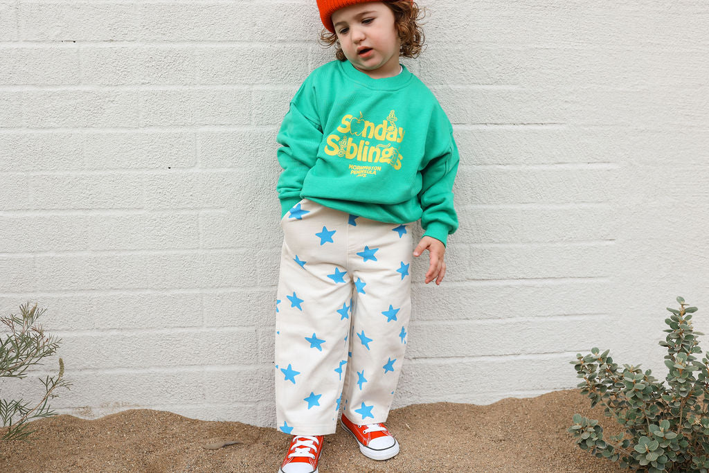 A young child stands against a light brick wall. They are wearing an oversized Doc Sweater Green by SUNDAY SIBLINGS made from French terry cotton with yellow text, cream-colored pants adorned with large blue stars, red sneakers, and a red beanie. Small green plants are visible in the foreground.