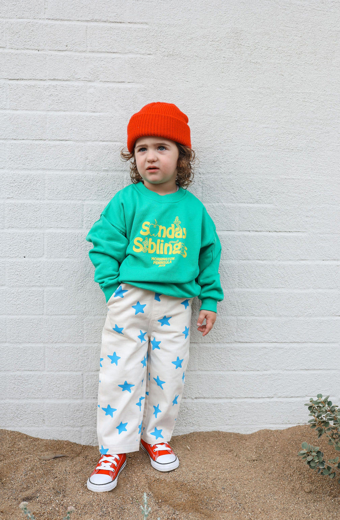 A young child stands against a white brick wall, wearing an oversized Doc Sweater Green made from French terry cotton with "SUNDAY SIBLINGS" written on it in yellow, white pants with blue stars, and red sneakers. The child has curly hair covered by a bright orange beanie and a neutral expression.