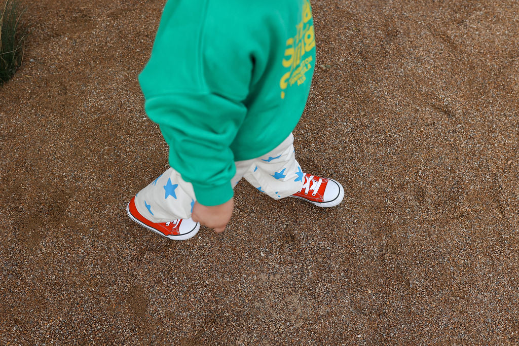 A child is walking outdoors on a gravel path, wearing the SUNDAY SIBLINGS Doc Sweater Green in an oversized fit, white pants with blue star patterns, and bright red sneakers. The focus is on the child's lower body, with the upper body partially visible.