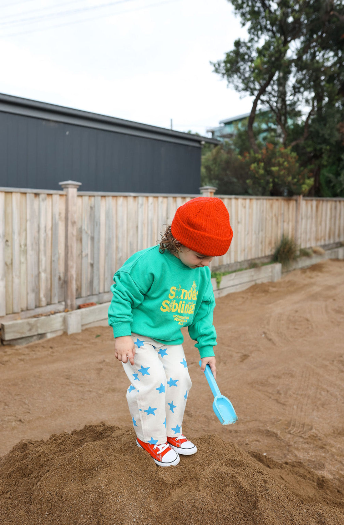 A young child wearing a bright red beanie, an oversized Doc Sweater Green from SUNDAY SIBLINGS made of soft French terry cotton, and white pants with blue stars stands on a dirt mound holding a blue shovel in one hand. The child is outdoors, with a wooden fence and trees in the background.