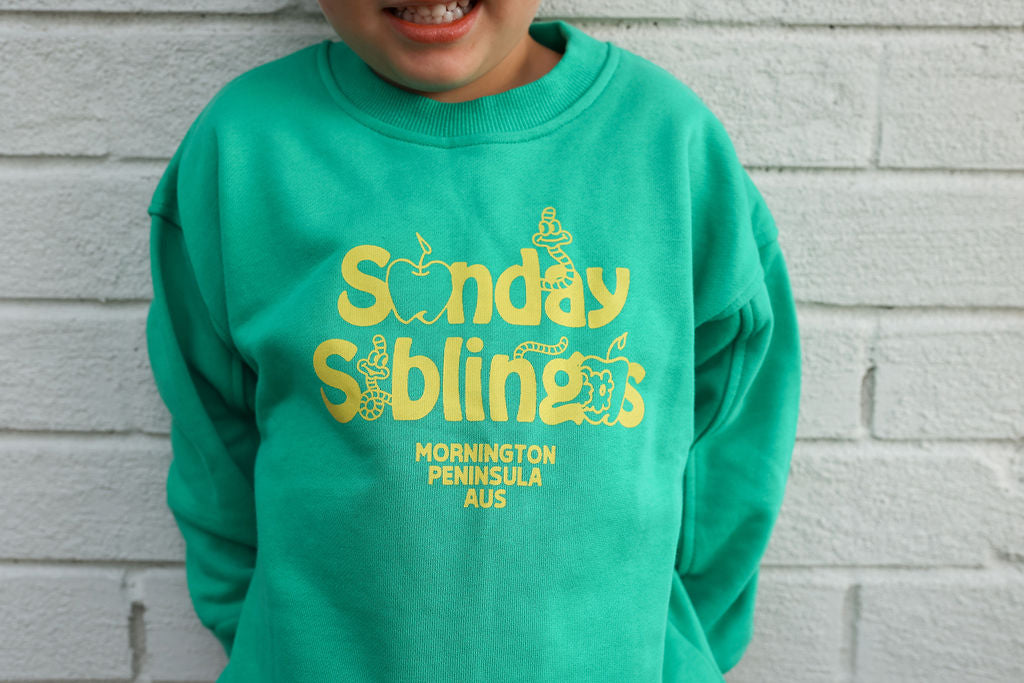 A child wearing an oversized fit, Doc Sweater Green from SUNDAY SIBLINGS, crafted from teal French terry cotton with yellow text that reads "Sunday Siblings" and "Mornington Peninsula AUS," adorned with apple and snake illustrations. The child stands against a white brick wall, smiling.