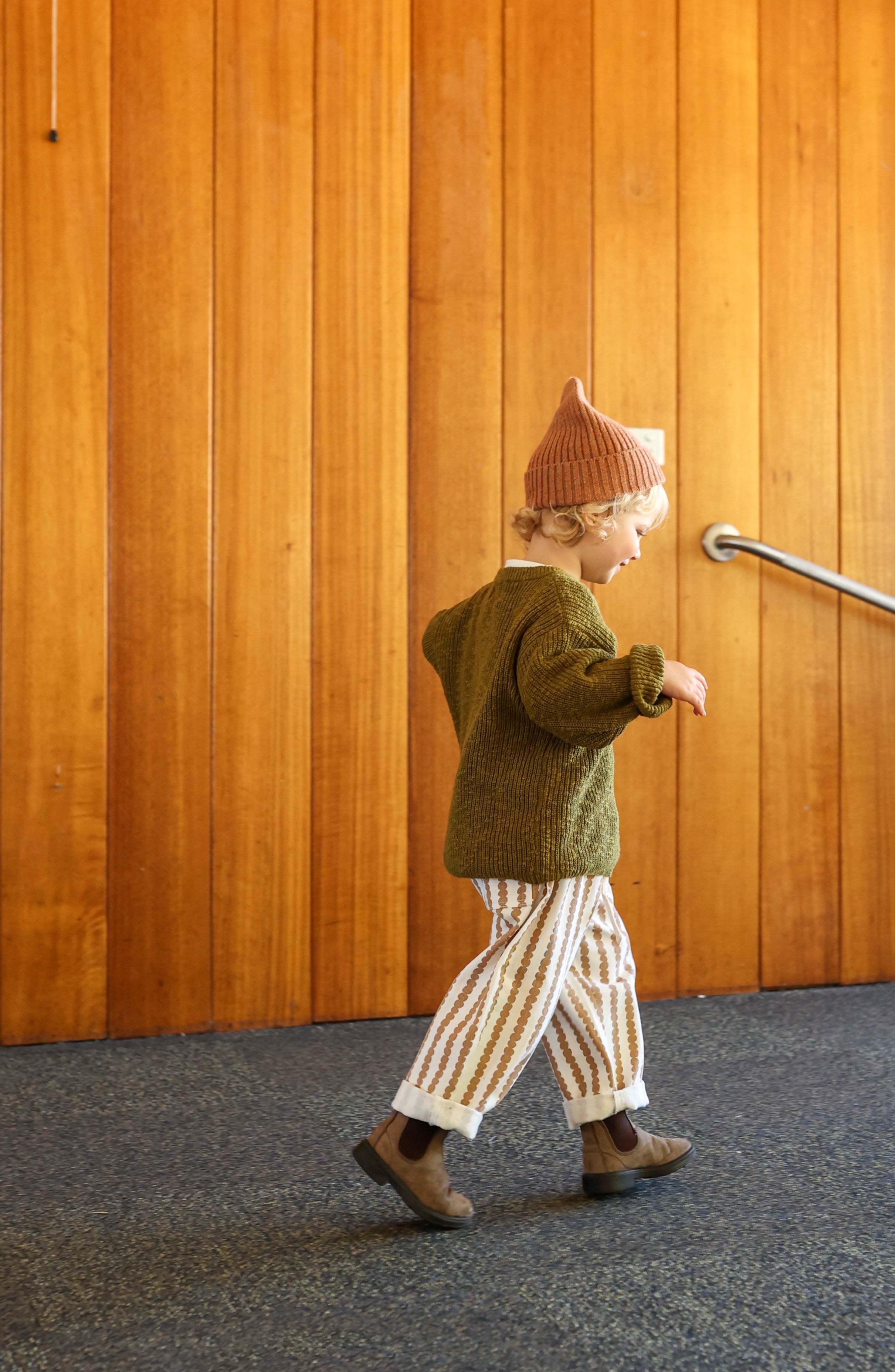 A child wearing a knit hat, green sweater, and GROWN Everyday Denim Jeans Stacked Pebble walks indoors with vertical wooden panel walls and a visible metal handrail.