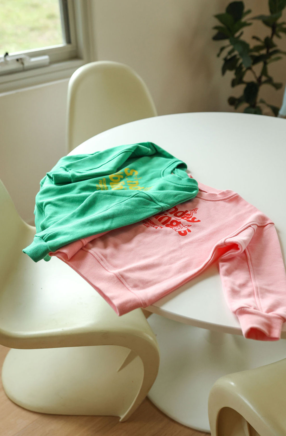 Two French terry cotton sweatshirts are laid out on a white round table with modern white chairs around it. One sweatshirt, the Doc Sweater Green by SUNDAY SIBLINGS, features a yellow design, and the other is pink with a red design. A window and a plant are in the background.