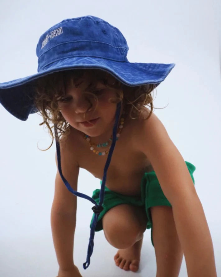 A child in a Ziggy Zaza ~ Everyday Scout Hat Blu by ZIGGY ZAZA crouches, wearing green shorts and a colorful beaded necklace. The wide-brimmed hat with an adjustable chin strap pops against the plain, light background.