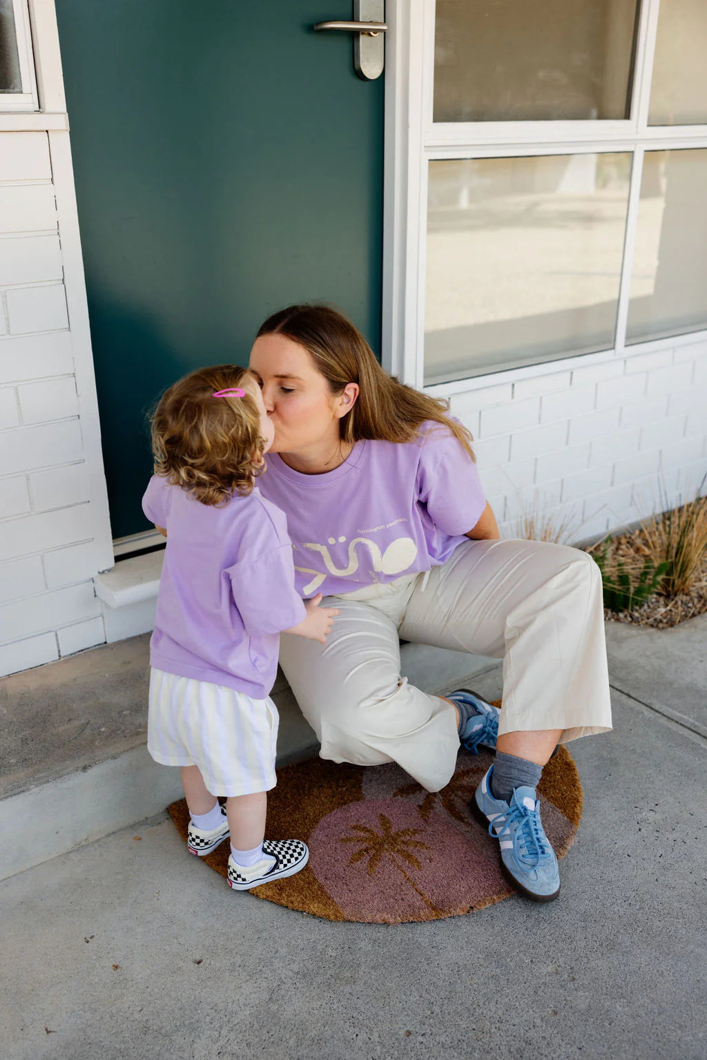 A woman on a doorstep, in an oversized Sunday Siblings Il Sole Tee Purple/Cream, kisses a toddler. Both wear matching shirts. She pairs hers with beige pants and blue sneakers, while the child has striped shorts and checkered shoes. They unwind on a palm-designed mat.