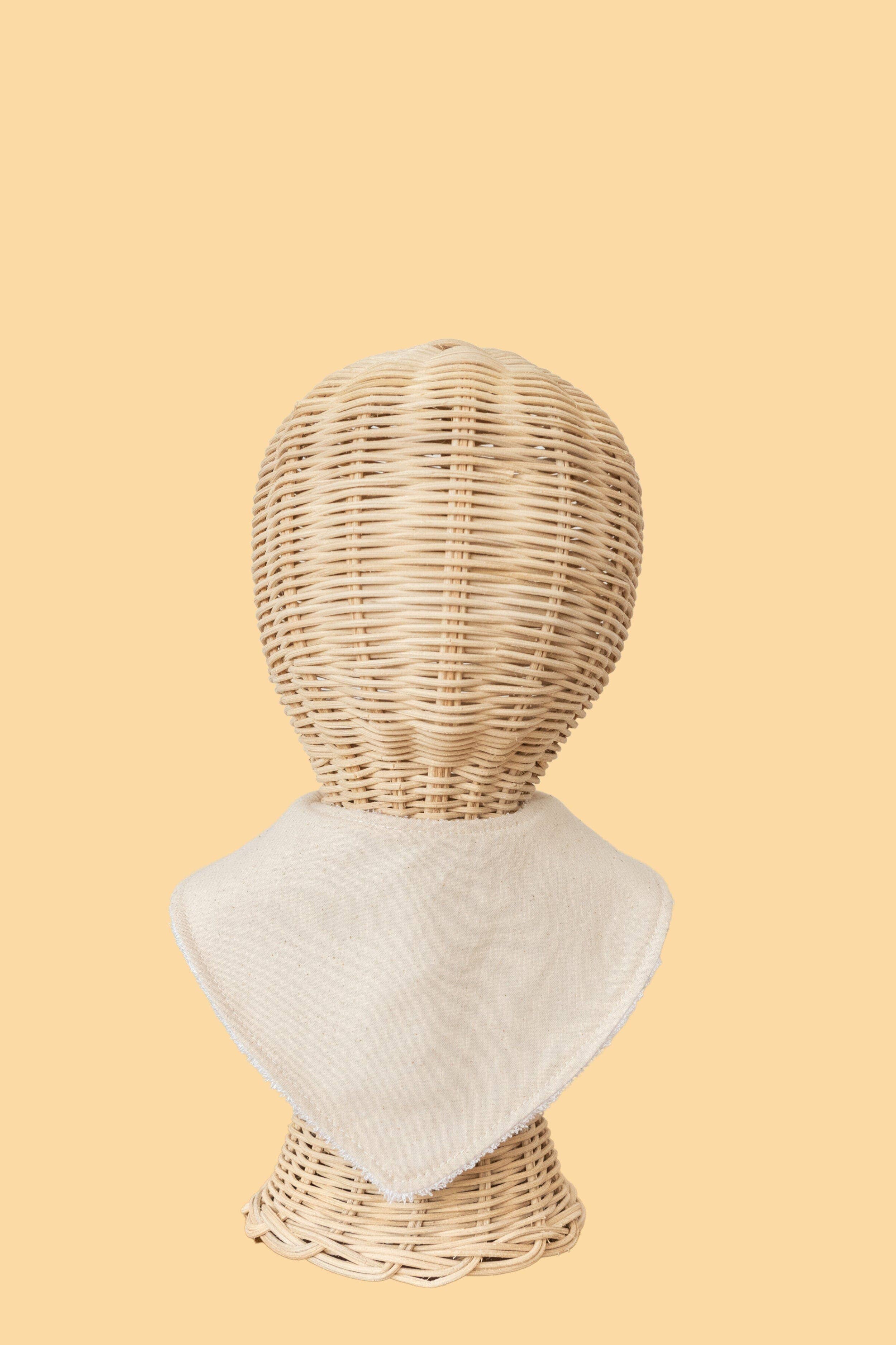 Against a plain beige backdrop, a wicker mannequin head is adorned with KIIN BABY's Dribble Bib Oat, crafted from white cloth.