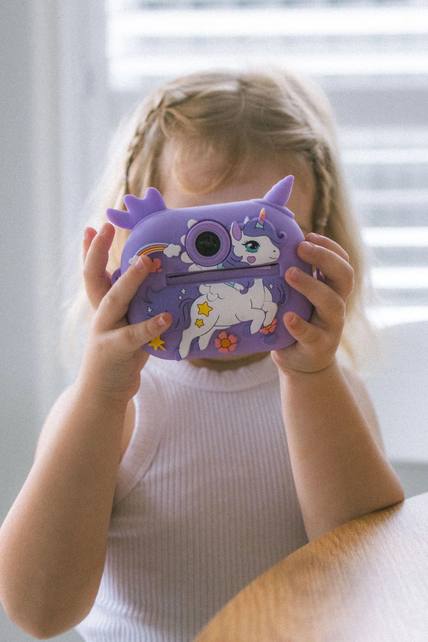 A child with braided hair holds an Oh My Frankie ~ Oh My Snap Instant Print Camera Purple with a unicorn design, sparking creativity. They sit at a wooden table, and white blinds are in the background.