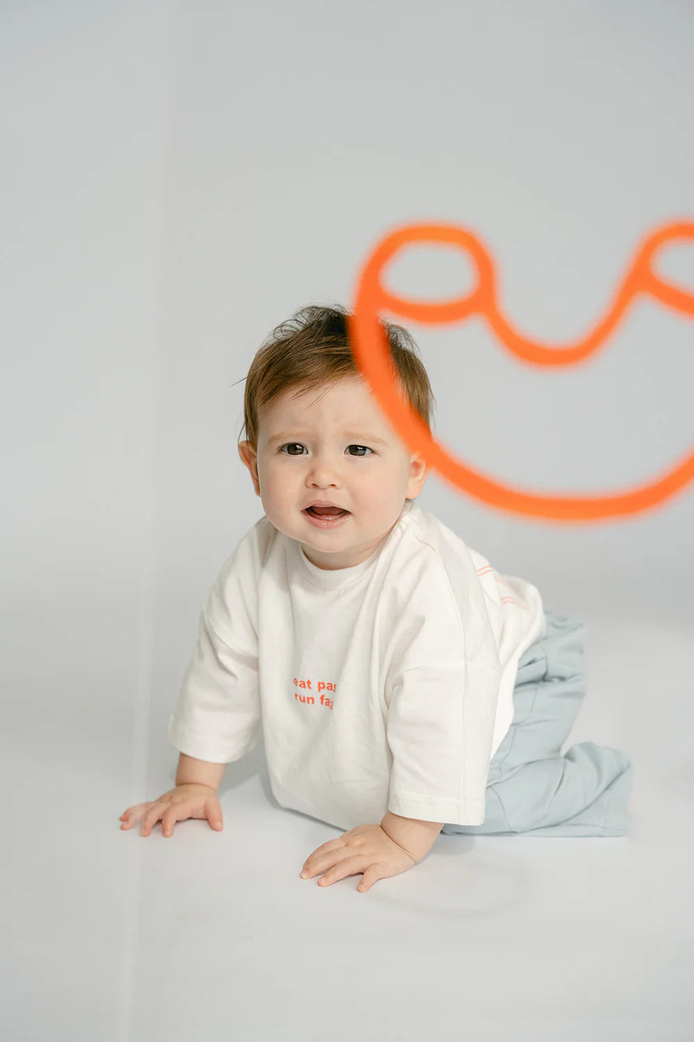 A baby dressed in an oversized fit white Pasta Tee from LENN LABEL and grey pants made of soft cotton jersey is crawling on the floor, looking curiously ahead. In the foreground, there's an orange squiggly smiley face drawn in the air, adding a playful element to the scene.