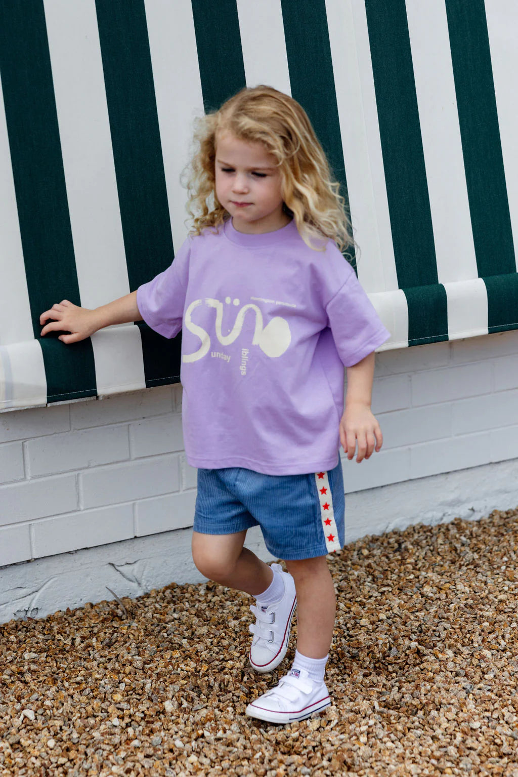 A young child with curly blonde hair walks on gravel wearing a SUNDAY SIBLINGS Il Sole Tee in Purple/Cream. They sport blue shorts with red stars and white sneakers, standing next to a building with a green and white striped awning.