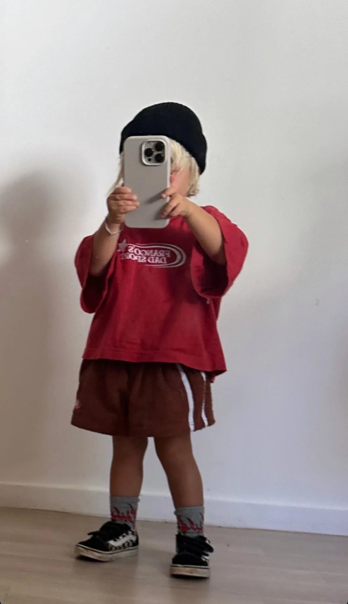 A young child stands against a plain wall in a red shirt, wearing FRANCO'S DAD PRE-ORDER Towel Shorts and a black beanie. With a large smartphone in hand, they capture the moment as their baggy fit sneakers peek from colorful socks—a playful summer staple ensemble.