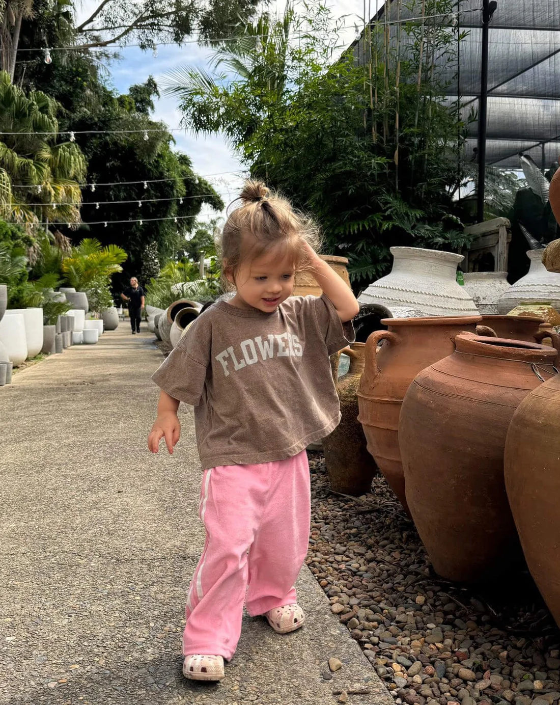 A young child in a brown "Flowers" T-shirt and ZIGGY ZAZA's Ziggy Zaza ~ Twin Stripe Terry Pant Pink strolls alongside large pottery. Touching their hair, they're surrounded by lush plants and potted trees. The 100% terry cotton fabric ensures comfort during playful adventures.