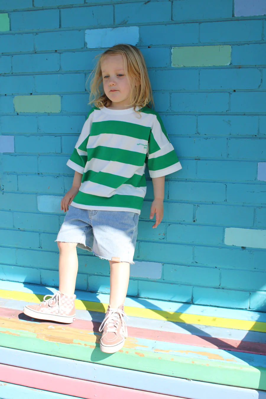 A child with long hair wearing the TWIN COLLECTIVE Twin Collective ~ Stripe Tee Green and denim shorts stands against a turquoise brick wall, leaning slightly with arms by their side. They wear high-top sneakers on a colorful striped surface.