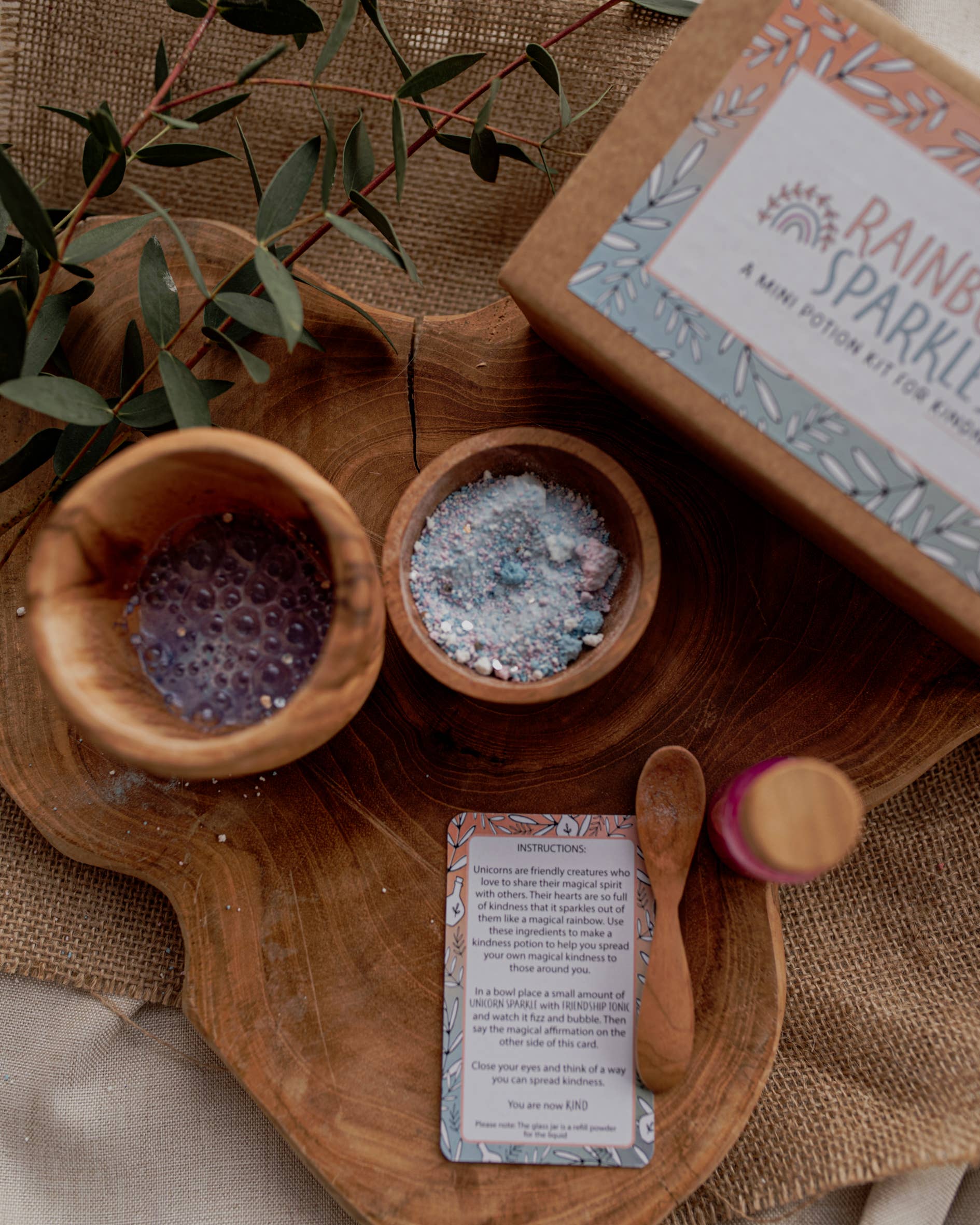 A wooden surface displays a small bowl of multi-colored bath salts, a larger cup with purple liquid, a wooden spoon, and a small bottle with a pink cap. A card with instructions hints at imaginative play. To the right, there's a cardboard box labeled "MINI Rainbow Sparkles Potion Kit (A potion for kindness)" by THE LITTLE POTION CO. Eucalyptus leaves complete this enchanting potion kit.