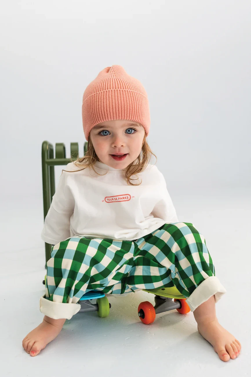 A young child with long hair wearing an orange knit beanie, a white long-sleeve shirt with a red logo, and SONNY LABEL Picnic Pocket Pants Forest, sits on a green toy skateboard. The child is barefoot and smiles at the camera against a white background.