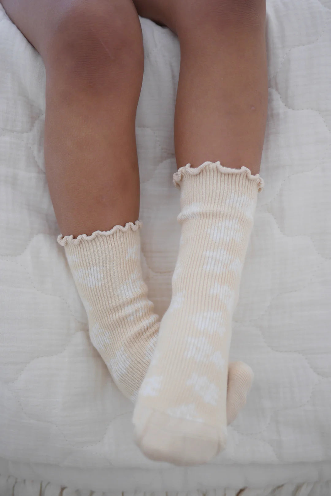 Close-up image of a child's legs and feet wearing ZIGGY LOU's Socks Banksy with white polka dots. The baby socks have ruffled tops and rest on a white quilted surface, highlighting medium-dark skin tones. These charming socks are part of the Banksy + Beech Spring Collections.