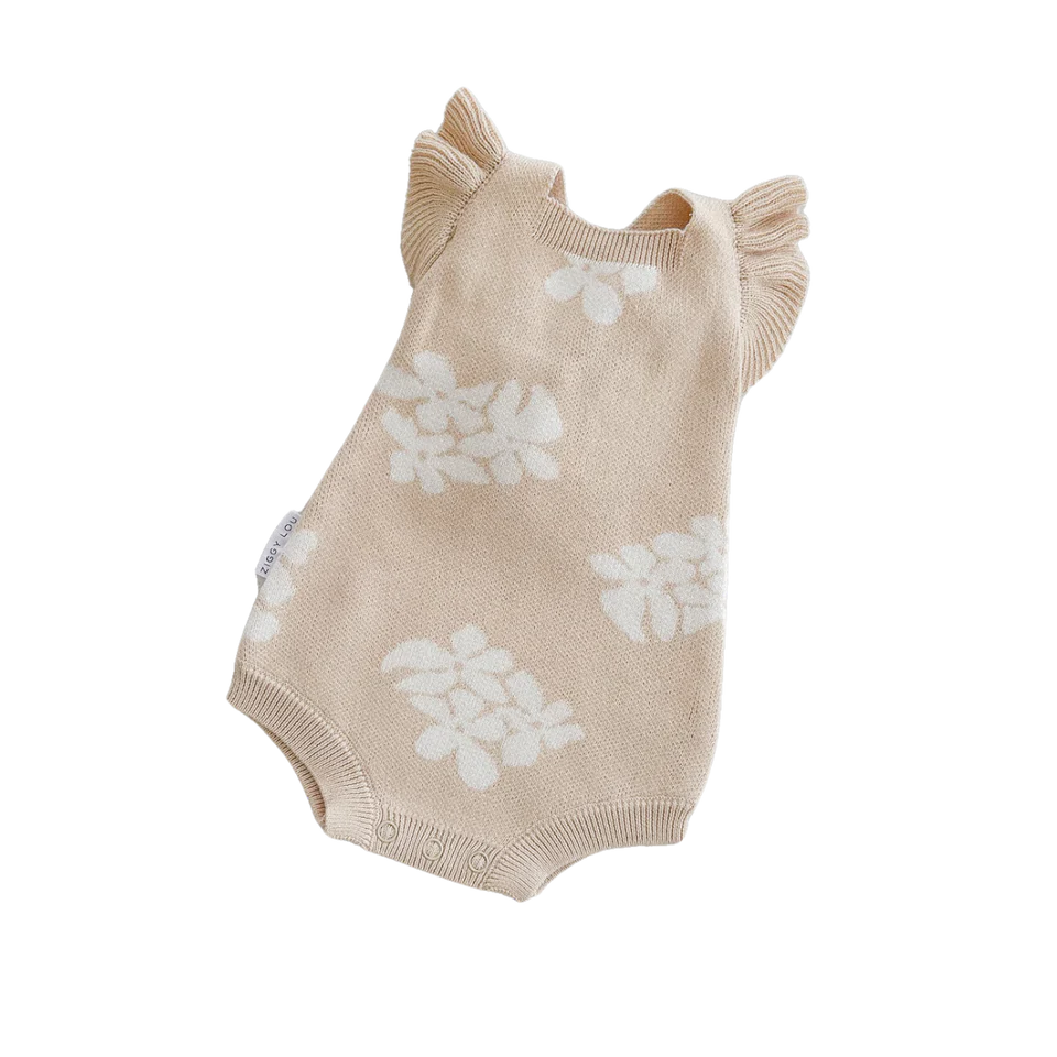 The Frill Romper Banksy from ZIGGY LOU is a beige, sleeveless knit romper adorned with white floral patterns. It features ruffled shoulder straps, ribbed leg openings, and functional snaps for easy changing. The soft, knitted material ensures it is suitable for a baby’s delicate skin.