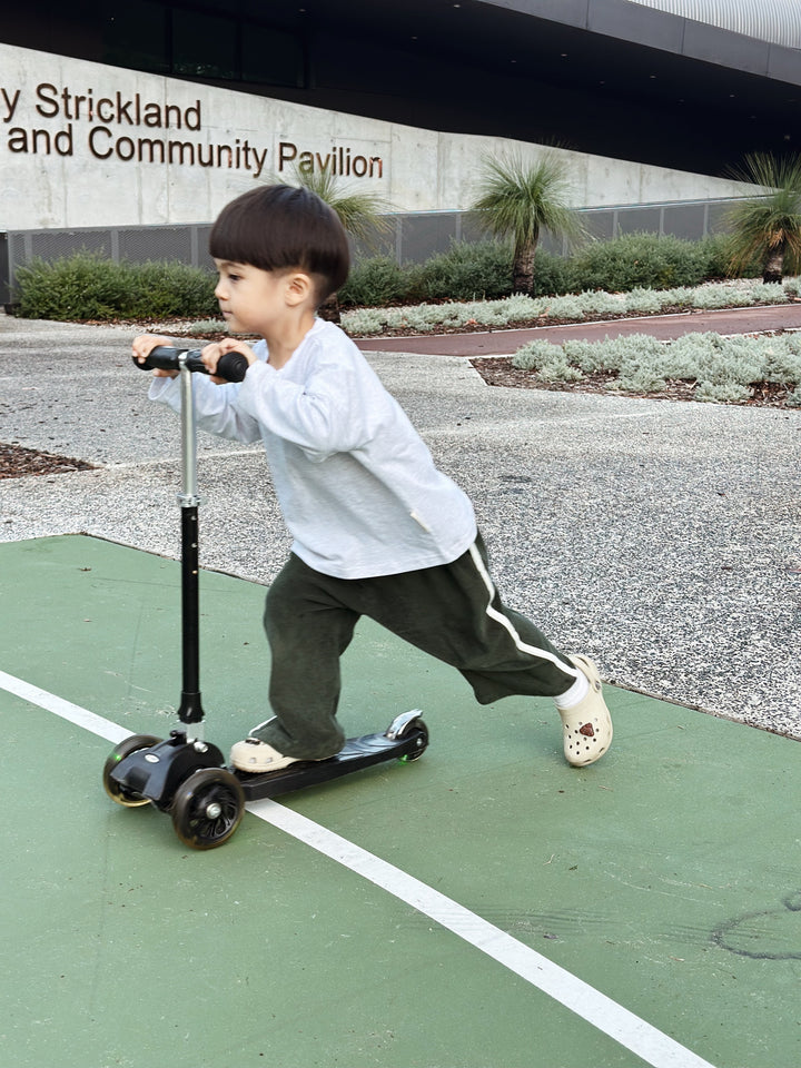 A young child wearing a long-sleeve shirt, TINY TROVE Darcy Racer Pants Moss with an elastic waistband, and crocs rides a three-wheeled scooter on a green surface outdoors, in front of the "Betty Strickland Community Pavilion." The area is landscaped with gravel, shrubs, and palm plants.