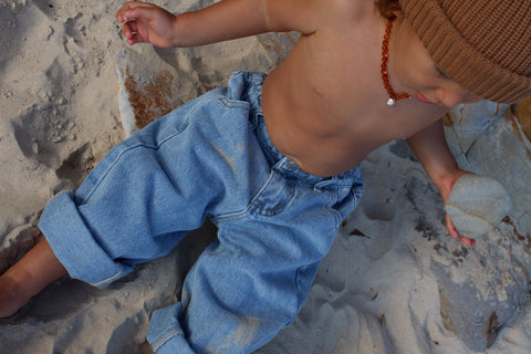 A child is sitting on a sandy surface, wearing light blue Limitless Jean Sky Blue Denim from SLEEPY DENIM with an adjustable waistband rolled up at the cuffs, a brown knit hat, and an amber necklace. The child is shirtless and holding a round stone in their right hand.