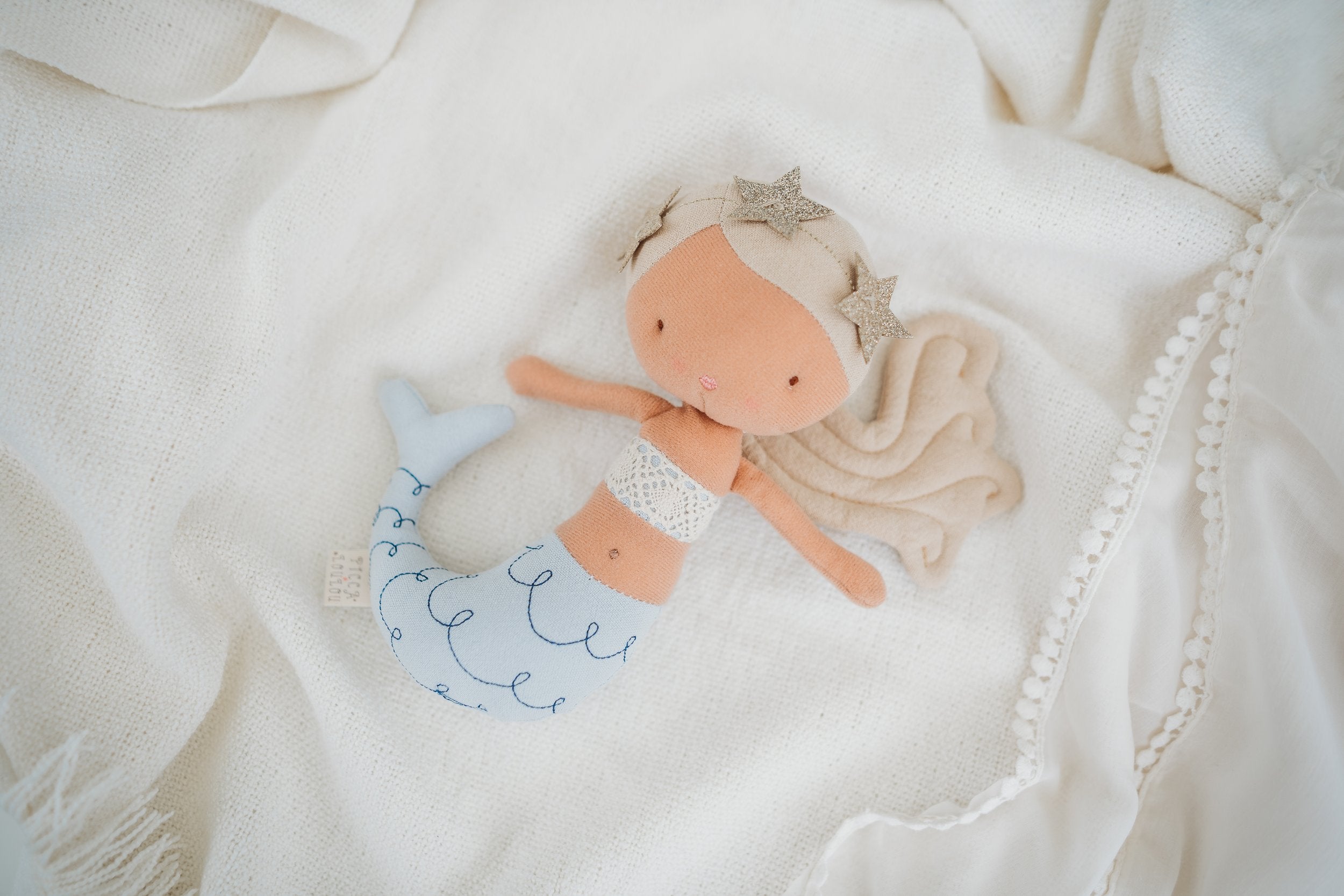 The PICCA LOULOU Mermaid Pearl 22cm doll is a soft, plush mermaid with light brown hair adorned with star-shaped clips. She features a light blue tail with wave patterns and wears a lace top, resting gracefully on a cream-colored fabric background. Perfectly crafted by Bon Ton Toys.