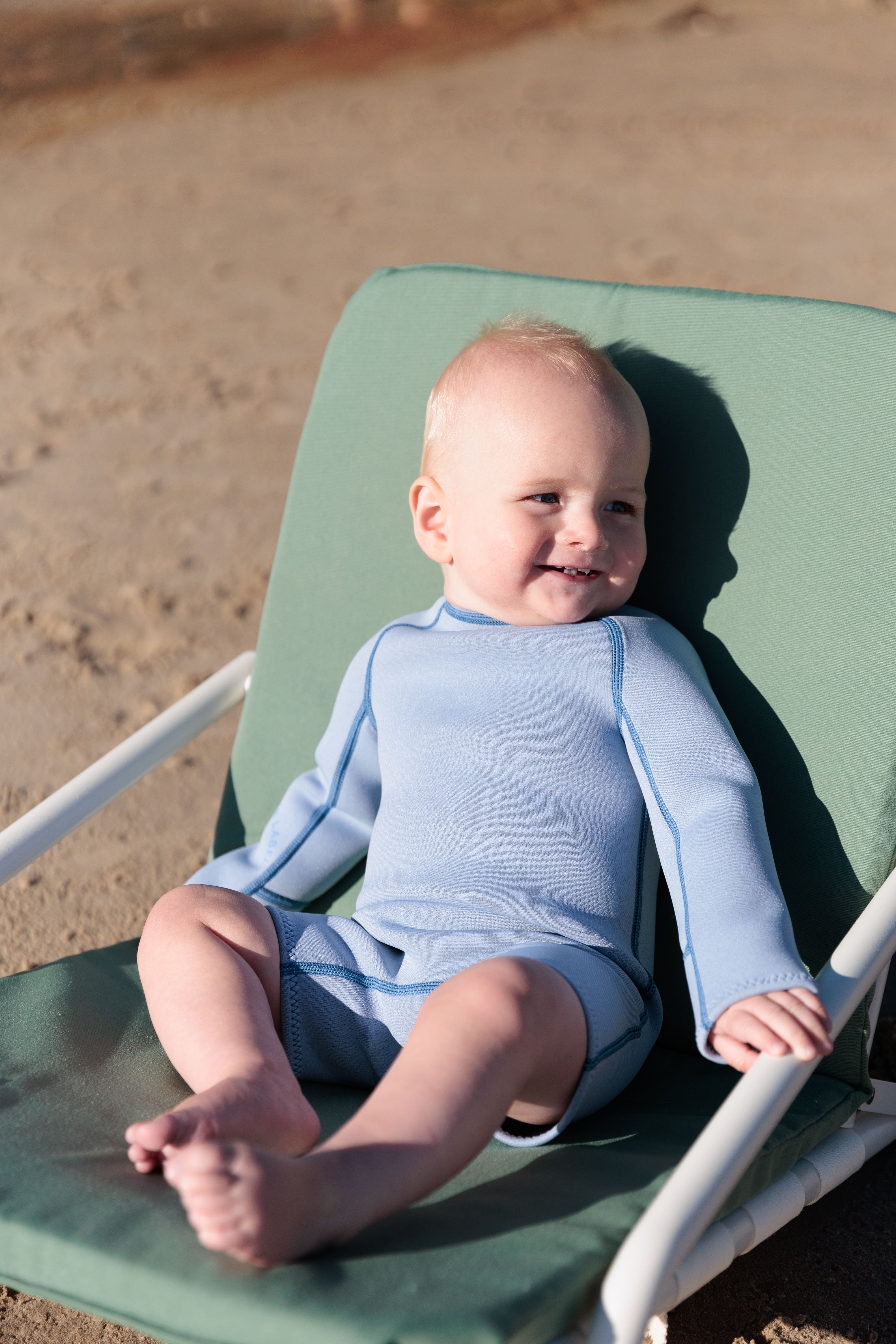 A baby dressed in a SUMMER SUN LABEL's Springsuit Wetsuit in Dusty Blue/Marine smiles and enjoys the sun with UV protection while relaxing on a green lounge chair at the sandy beach.