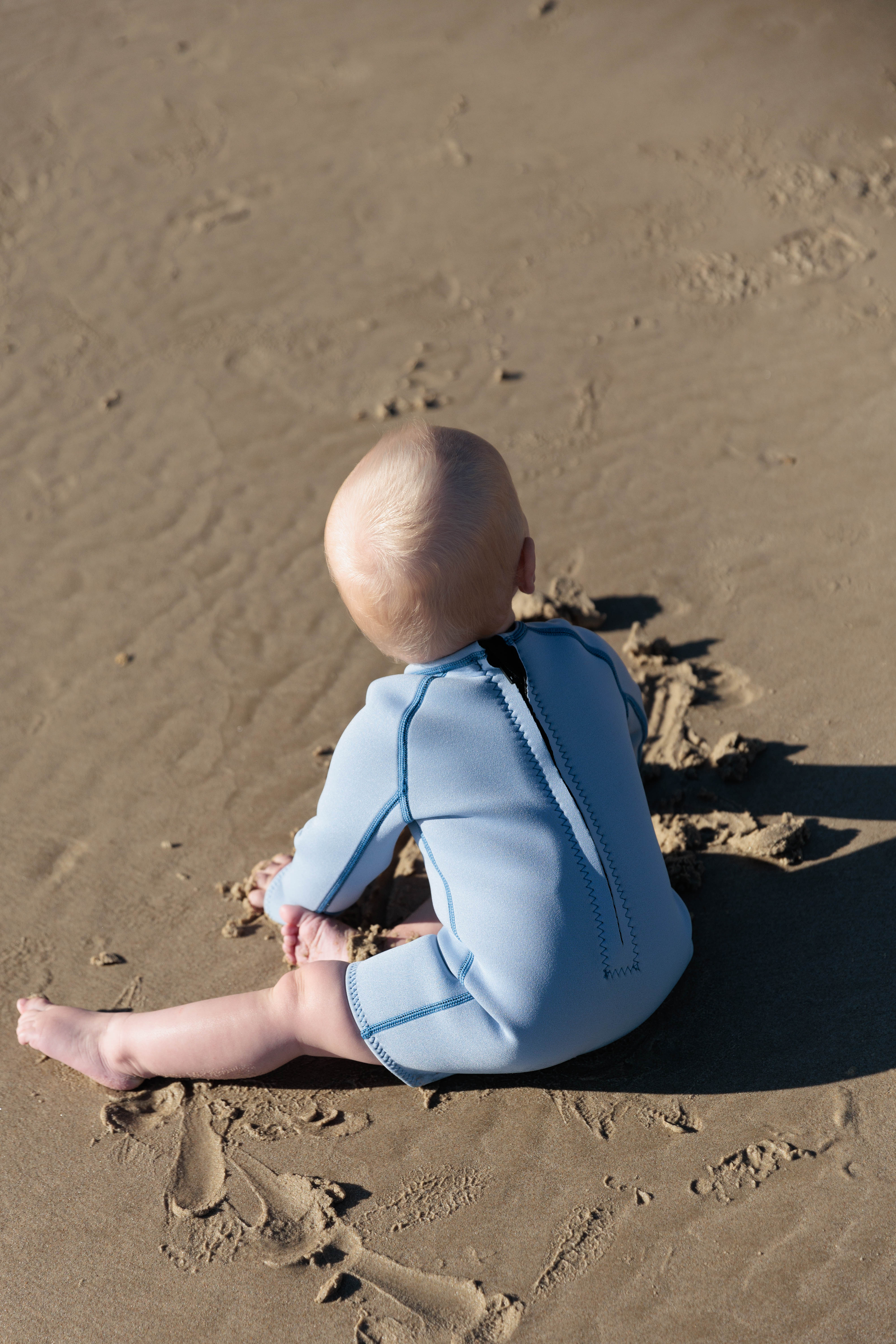 A baby wearing the Long Sleeve Springsuit Wetsuit in Dusty Blue/Marine by SUMMER SUN LABEL sits on a sandy beach, facing away. The sun casts a shadow on the sand, which is adorned with small footprints and natural textured patterns that echo nature's own contrast collection.