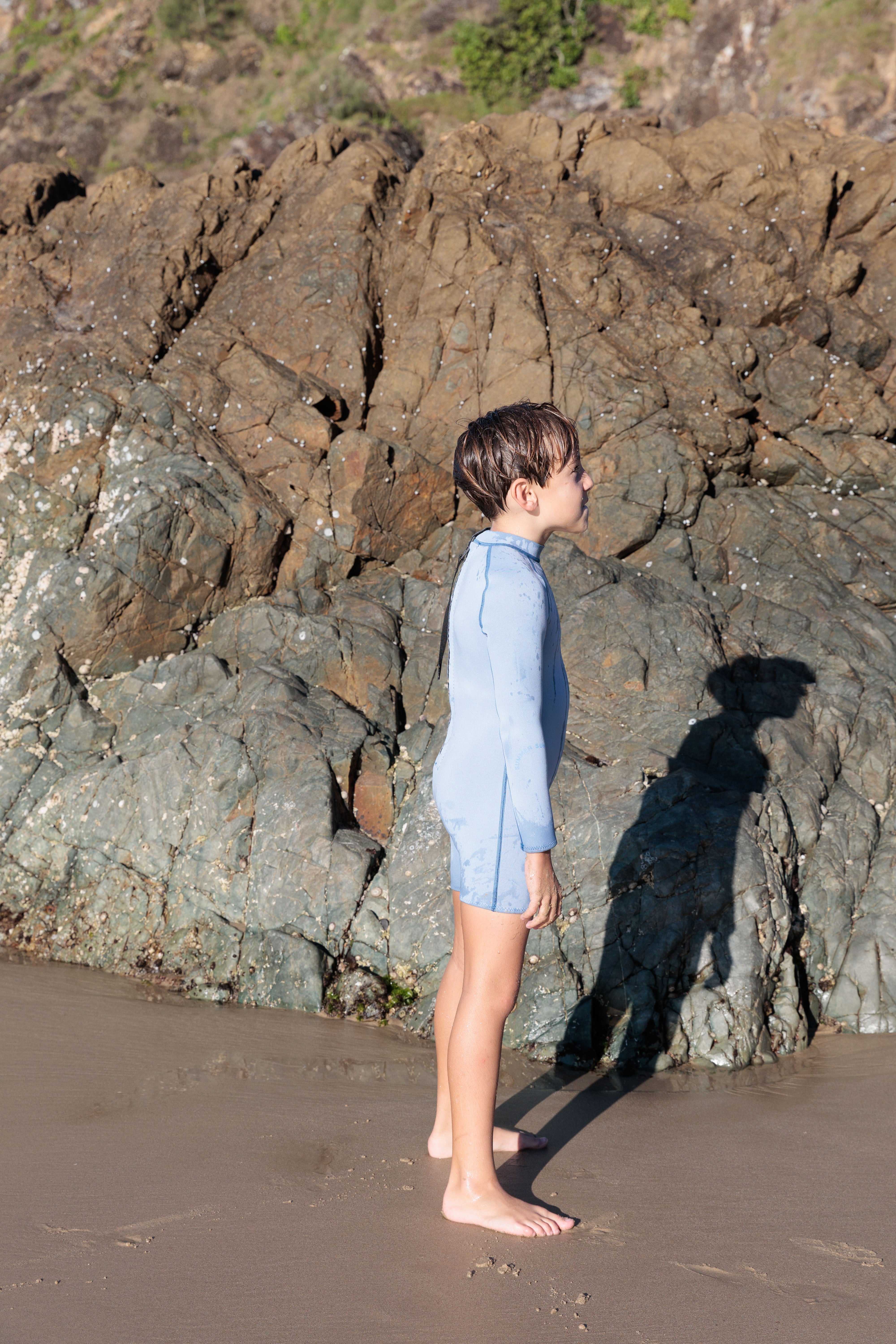 A child wearing a SUMMER SUN LABEL Long Sleeve Springsuit Wetsuit in Dusty Blue/Marine stands on a sandy beach, gazing at the towering rocky cliffs. The sun casts a long shadow on the sand, accentuating their attire from the Contrast Collection made of Yulex Foam material.