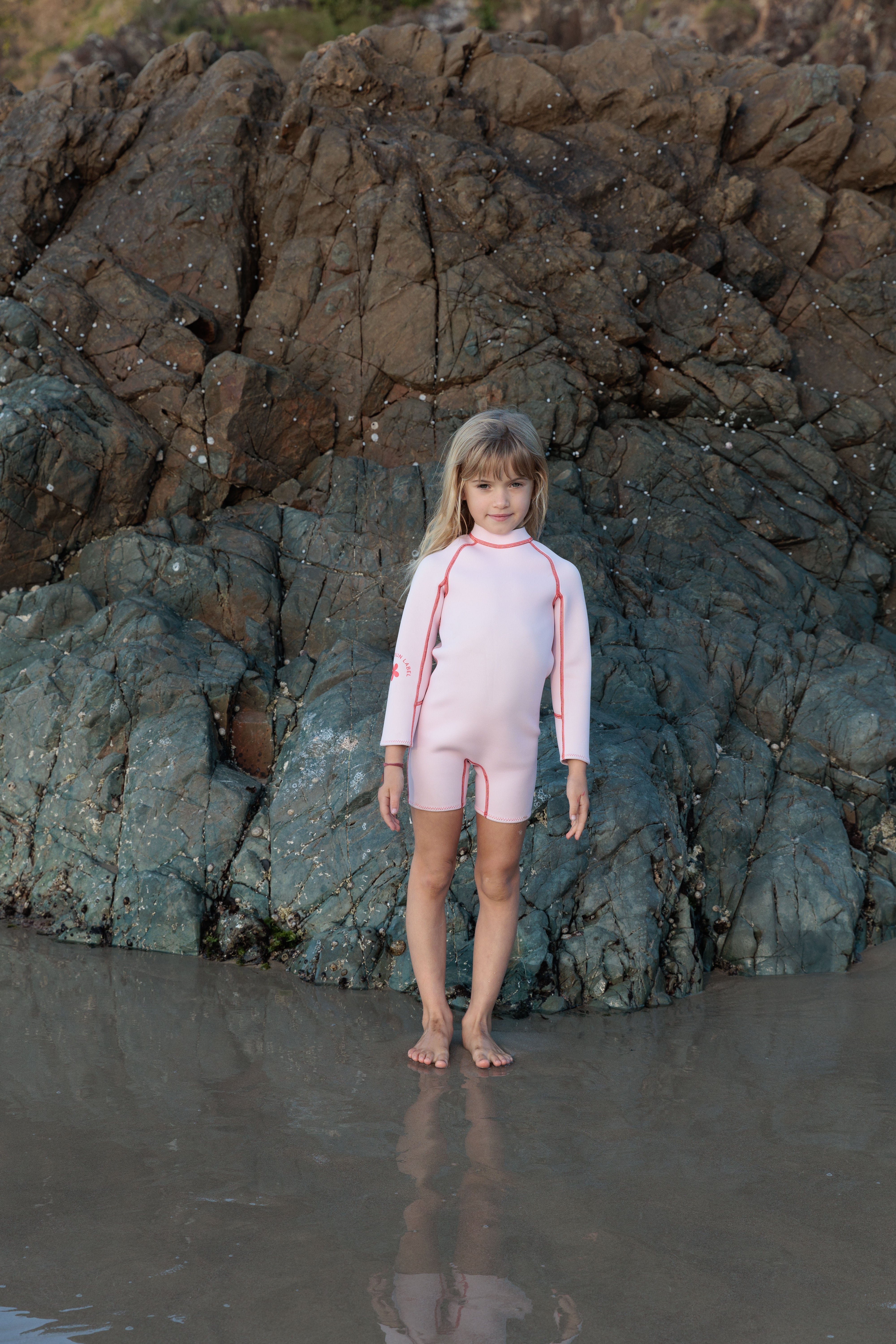 A young girl stands barefoot on wet sand with a rocky backdrop, wearing the Long Sleeve Springsuit Wetsuit in Powder Pink/Tomato from SUMMER SUN LABEL, crafted from sustainably sourced natural rubber. The seaweed-covered rocks and her reflection in the shallow water enhance the beach's natural ambiance.