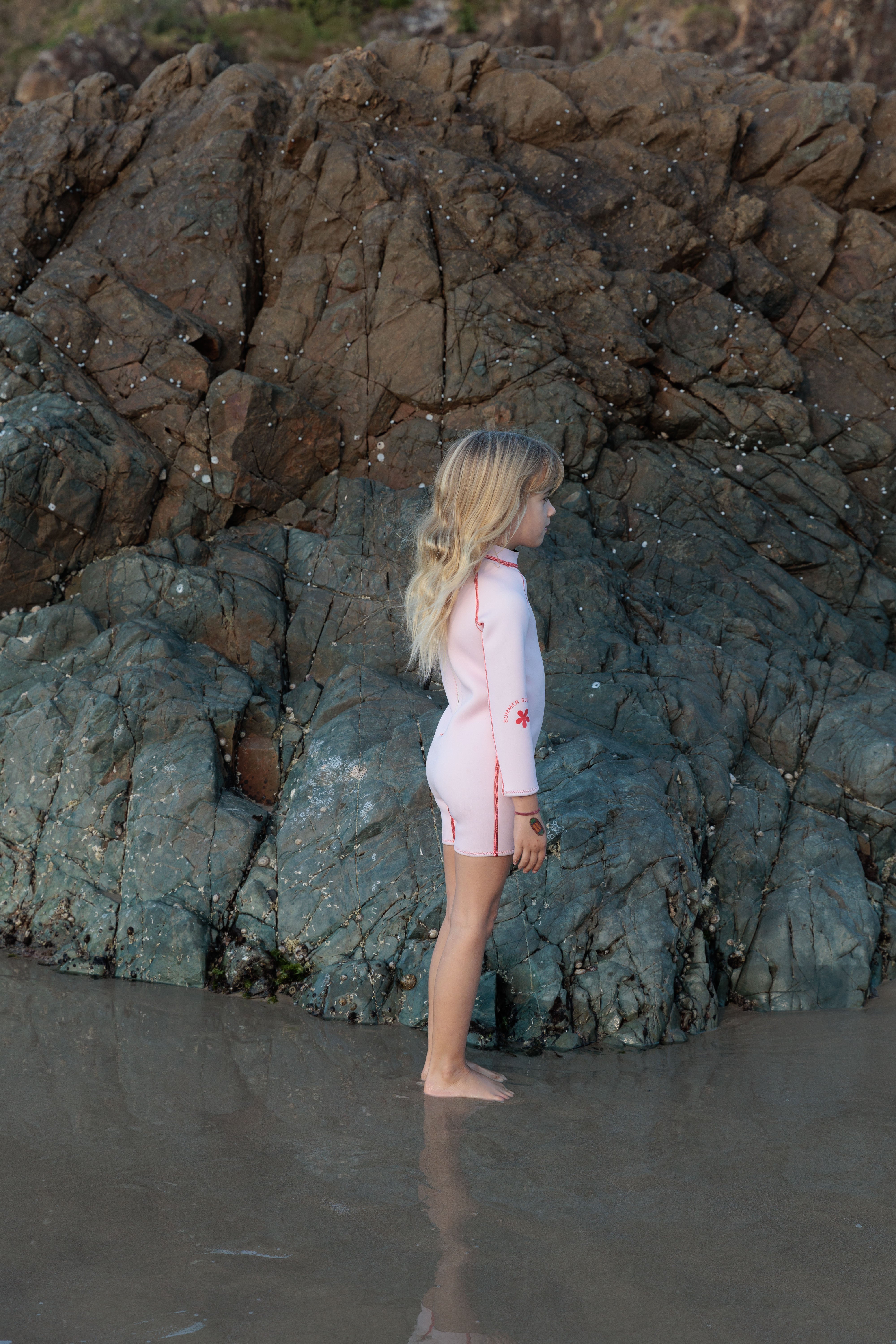 A child with long hair stands barefoot on the wet sand near rocky terrain, wearing the SUMMER SUN LABEL's Long Sleeve Springsuit Wetsuit in Powder Pink/Tomato, crafted from sustainably sourced natural rubber. The rocks display a textured blend of dark and light shades, completing a serene and natural scene.