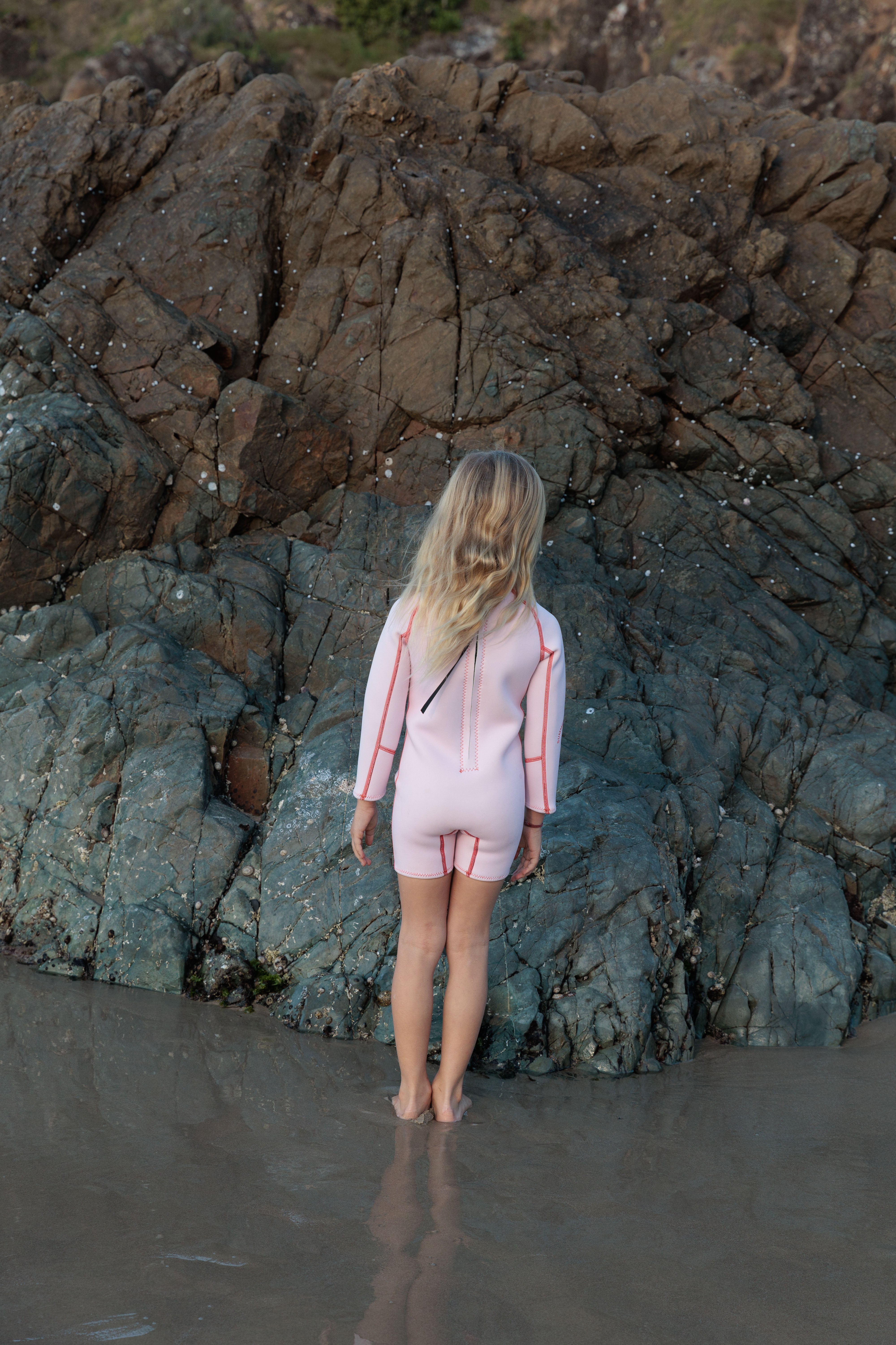 A child wearing the SUMMER SUN LABEL Long Sleeve Springsuit Wetsuit in Powder Pink/Tomato, made from sustainably sourced natural rubber, stands barefoot on wet sand with their long blond hair cascading down their back, gazing at a rocky cliff. The scene is set outdoors by the water, where textured rocks are partially adorned with seaweed or barnacles.