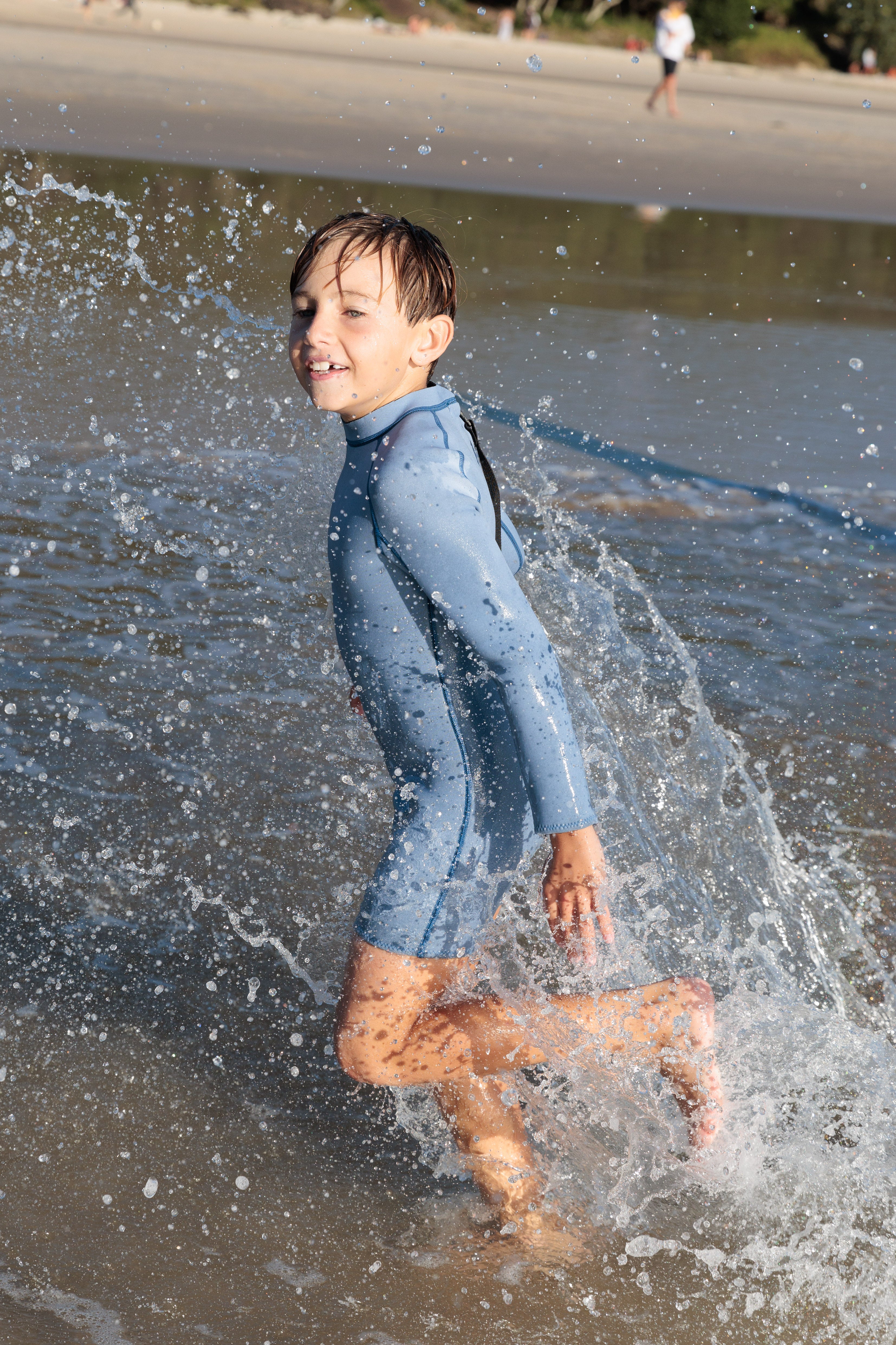 A child joyfully runs through shallow ocean waves at the beach, wearing a Springsuit Wetsuit in Dusty Blue/Marine from the SUMMER SUN LABEL. Splashes of water surround the child, with a sandy shoreline and trees visible in the background.