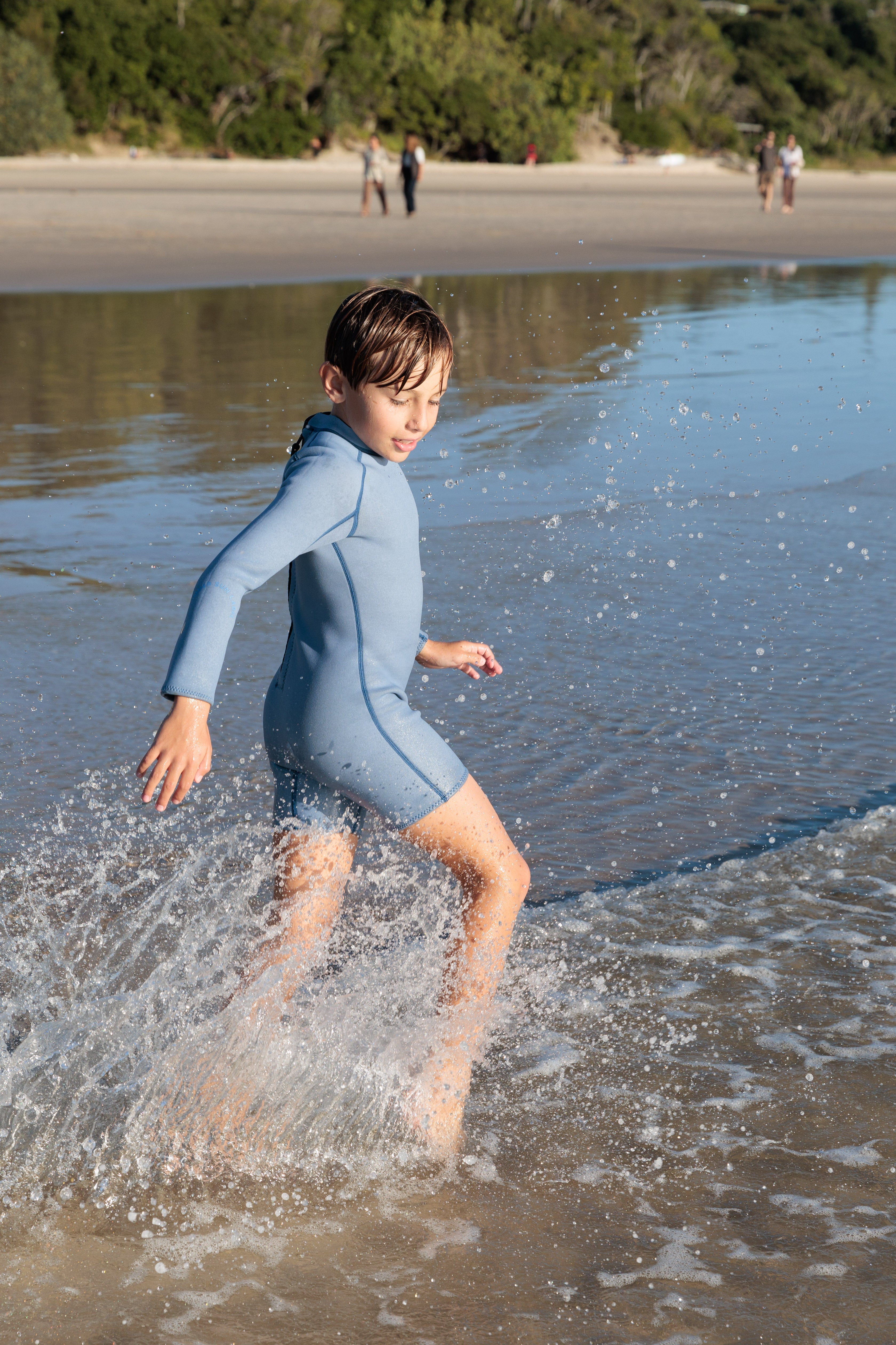 A young boy in a dusty blue and marine Long Sleeve Springsuit Wetsuit from SUMMER SUN LABEL runs through shallow ocean waves at the beach. The sun is shining, with trees and a few people in the background on the sandy shore.