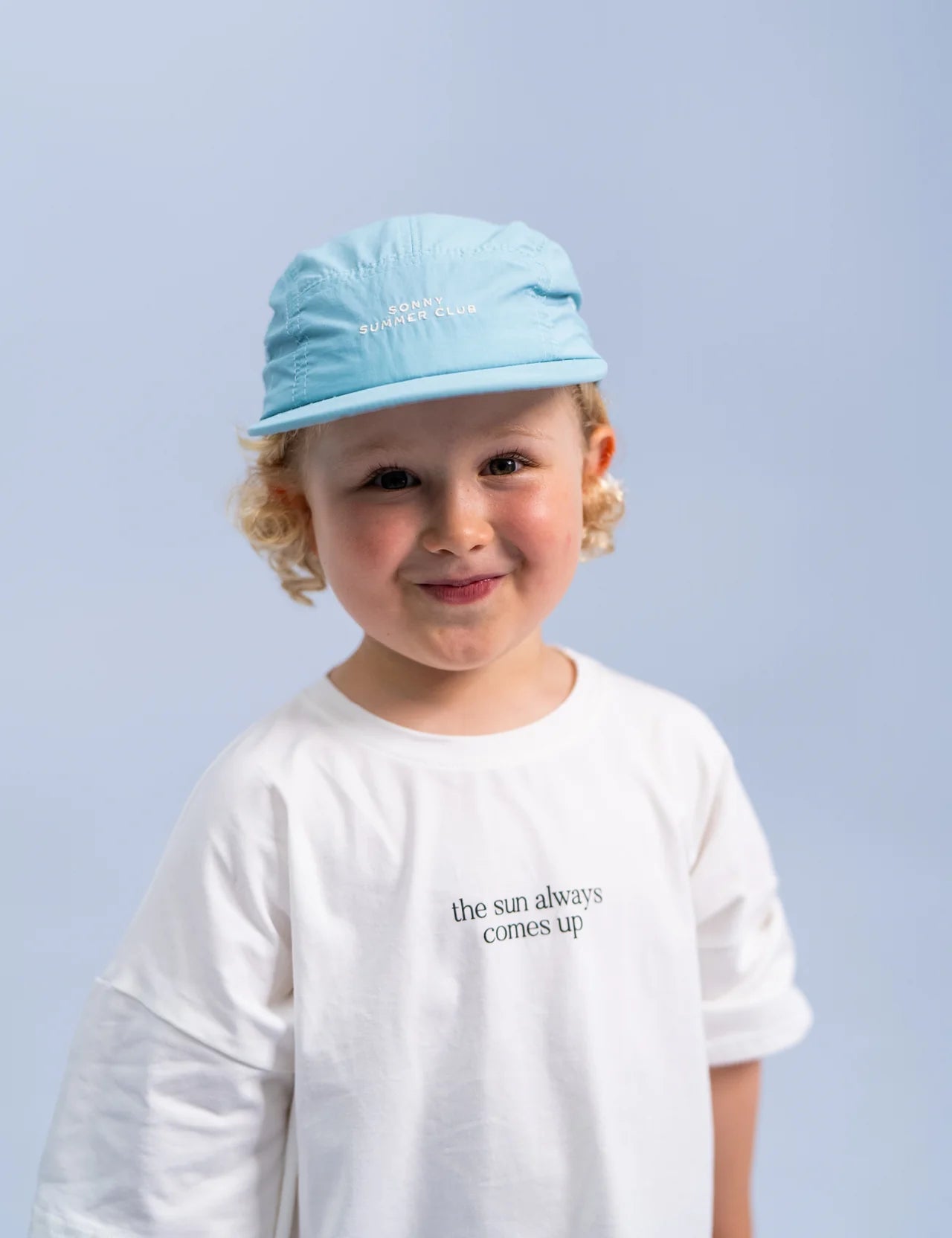 A young child with curly hair smiles while wearing the Summer Club Cap in Ocean by SONNY LABEL, along with a white T-shirt that says "the sun always comes up." The background is a soft blue.