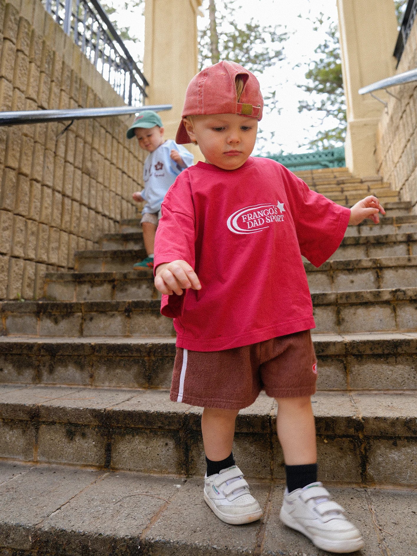 A toddler dressed in vibrant PRE-ORDER Towel Shorts by FRANCO'S DAD, a summer must-have, carefully navigates stone steps. Another child in a light blue shirt and green cap follows closely. The scene unfolds outdoors, framed by a brick wall and metal railings, capturing the essence of carefree childhood adventures.