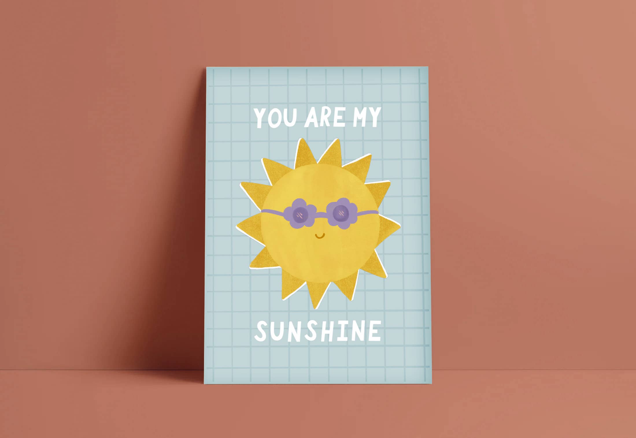 An art print with a picture of a sun wearing purple flower shaped sunglasses with the words "you are my" above it and the word "sunshine" below it. The print is on a blue background with a square pattern over the top. The art print is by Lauren Sissons Studio.