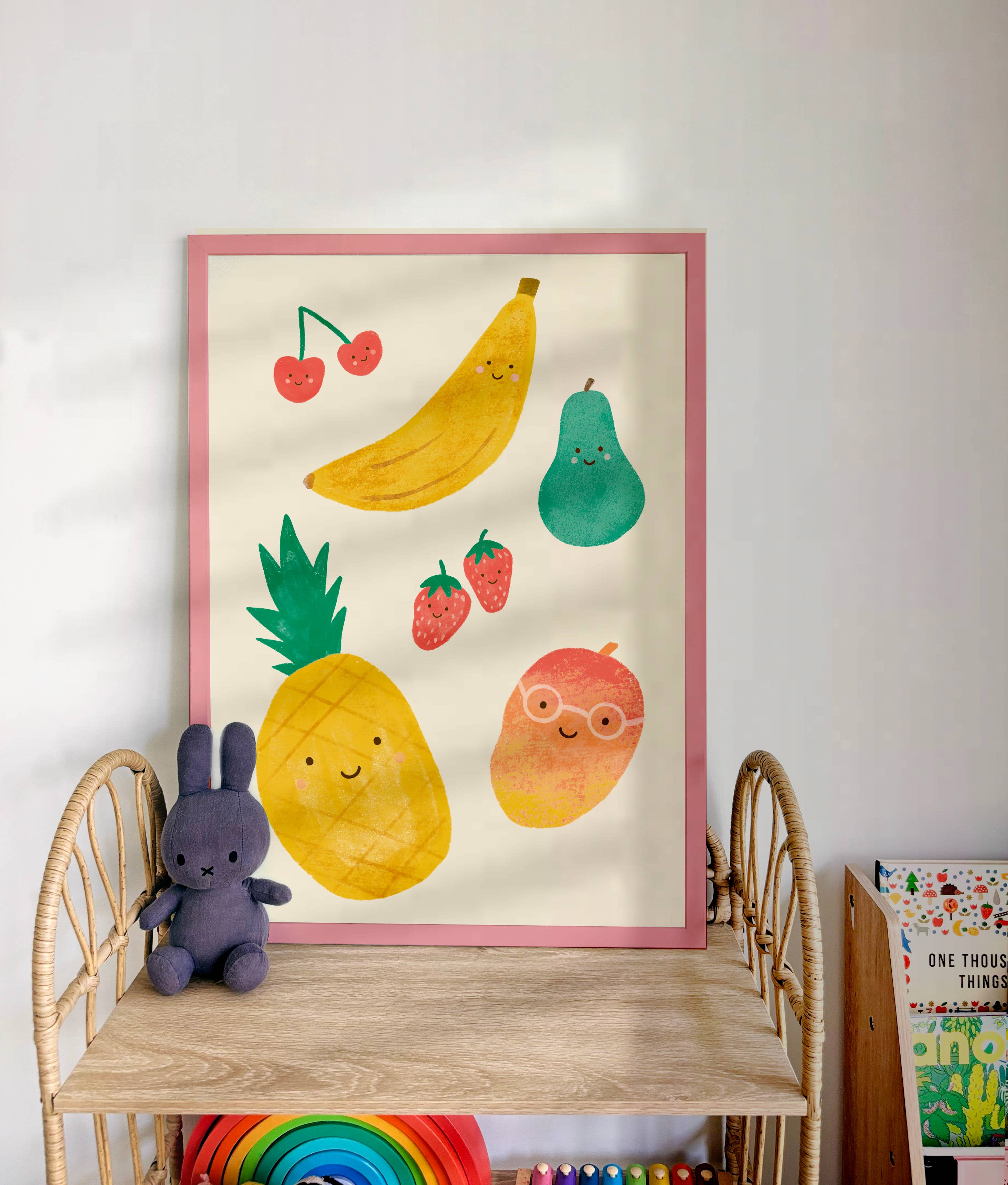 A wicker shelf holds a stuffed bunny toy and a row of children's books. Above the shelf is the LAUREN SISSONS STUDIO's Cute Fruit Wall Art Print, featuring cheerful illustrations of various smiling fruits such as bananas, pineapples, pears, mangoes, cherries, and strawberries—perfect for kid's room decor.