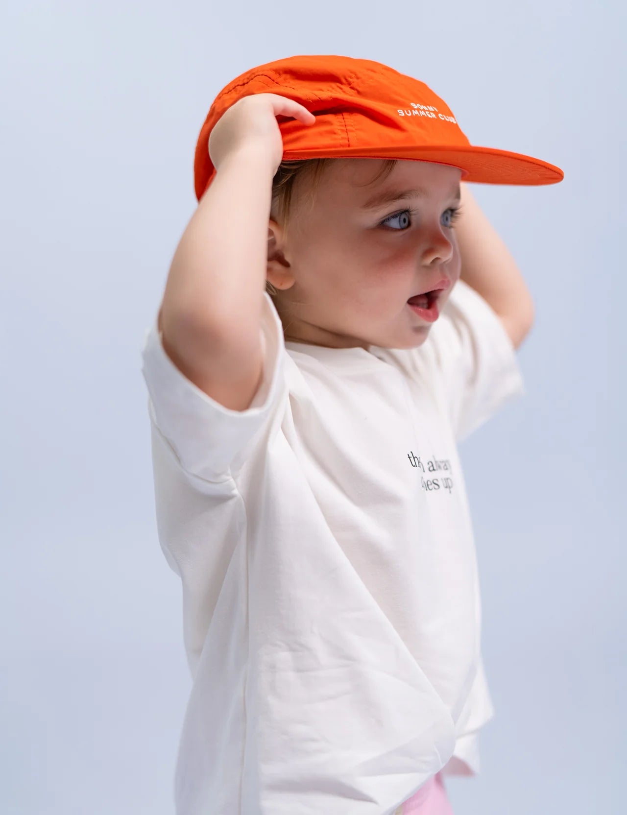 A toddler wearing a white T-shirt and the Summer Club Cap : Poppy by SONNY LABEL looks surprised or curious. The cap is orange, made of nylon, and features an adjustable velcro strap with white text on the front. The child has both hands on the cap against a plain, light background.