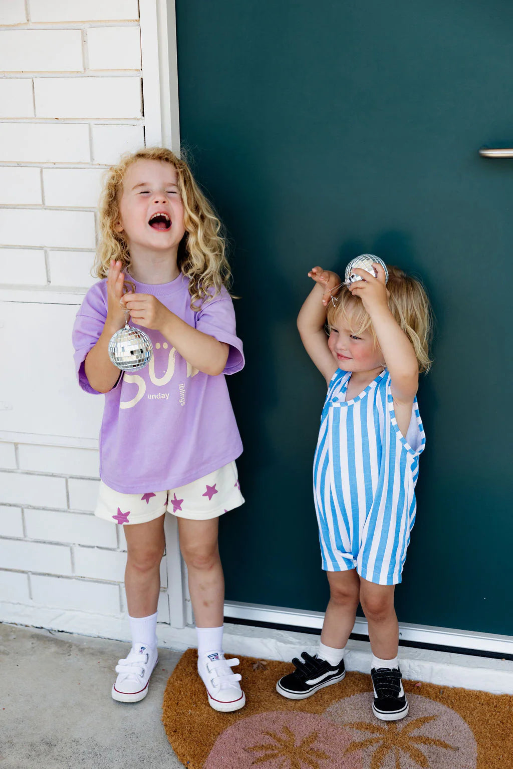 Two young children stand in front of a door. The child on the left is laughing, wearing Sunday Siblings ~ Kiddo Shorts Purple and a purple shirt with a star print. The child on the right is smiling in a cozy blue and white striped outfit, holding a shiny ornament.