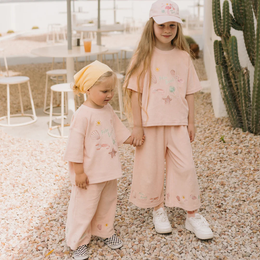 Two young children in matching pastel pink outfits stand holding hands outdoors. The older child sports a pair of Palm Beach Wide Leg Pants Terry Towel in Palm Flamingo Pink by GOLDEN CHILDREN, complemented by a hat and sneakers, while the younger child wears a yellow headscarf and checkerboard shoes. They are surrounded by white furniture and cacti.