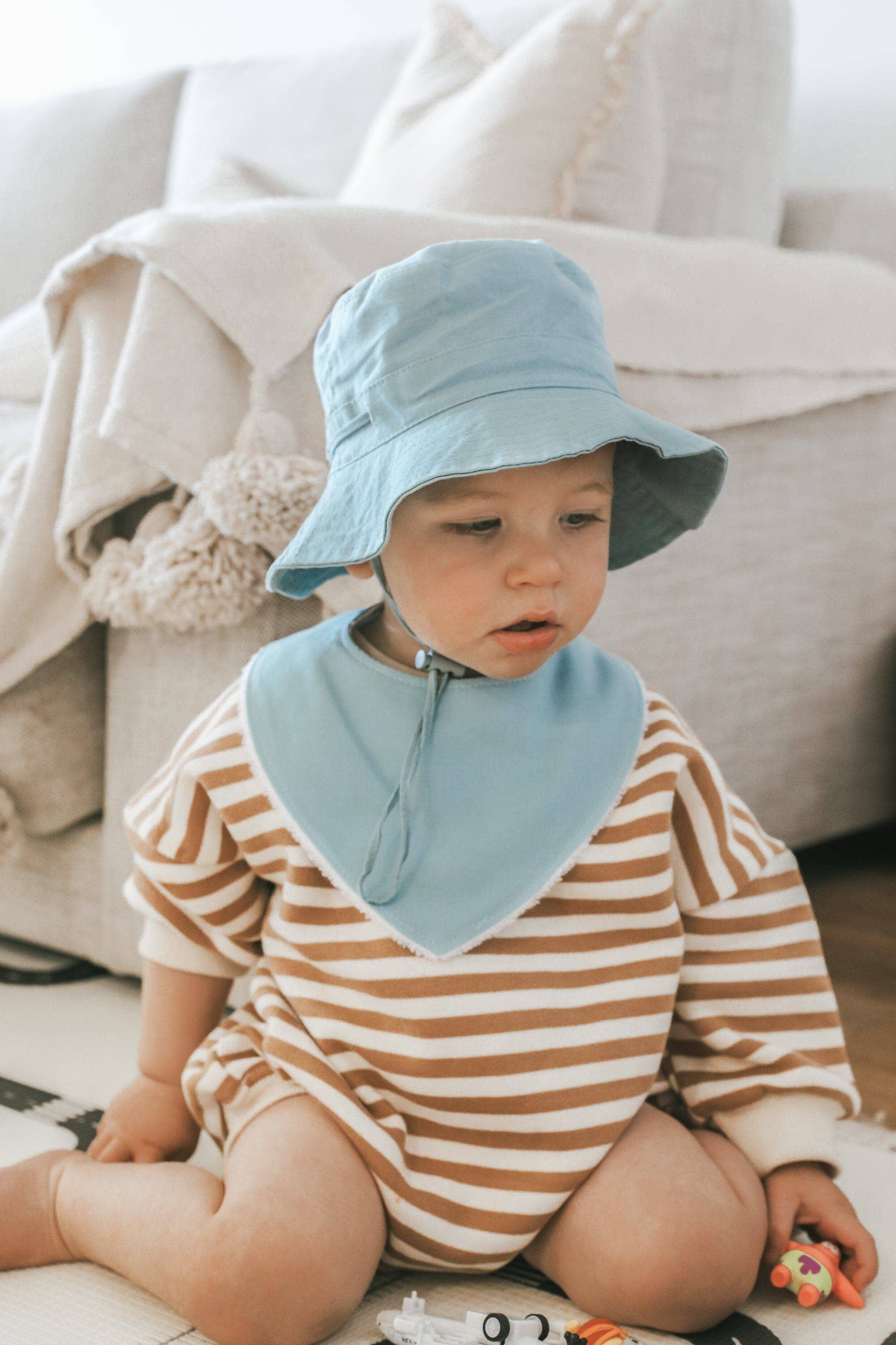 A baby sits on the floor wearing a blue sun hat and striped brown and white outfit, accompanied by Kiin Baby's absorbent bamboo/cotton Dribble Bib. Surrounded by toys, the scene features a beige sofa with a blanket in the background.